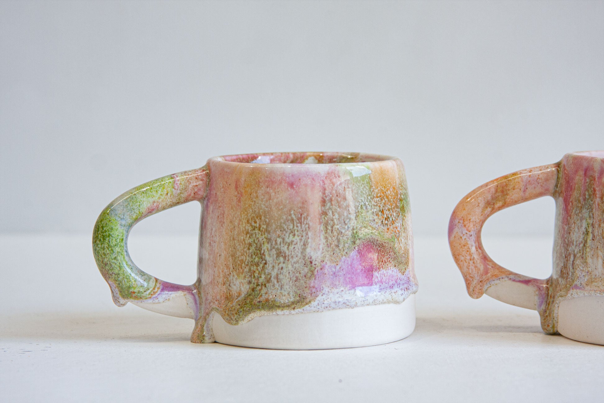 A handmade ceramic mug with a dripping glaze finish in pink, green, and gold tones, shown next to another partially visible mug on a light gray background.