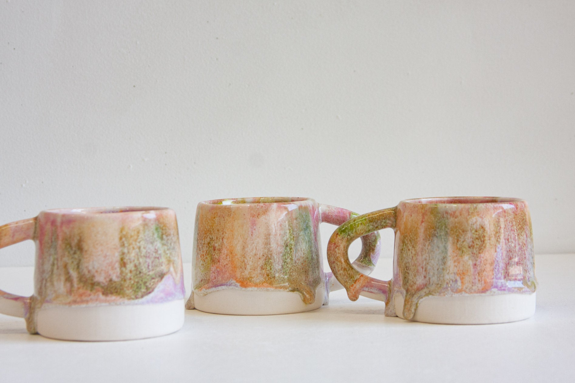 Three handmade ceramic mugs with handles, featuring a multicolor glaze with pink, green, orange, and brown tones dripping over a white glazed base, arranged on a white surface against a light gray background.