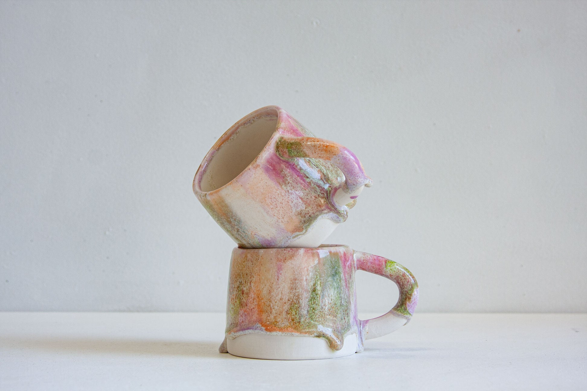 Two small handmade ceramic mugs stacked on top of each other, featuring a marbled, dripping glaze with pink, green, orange, and white tones, set against a plain light gray background.