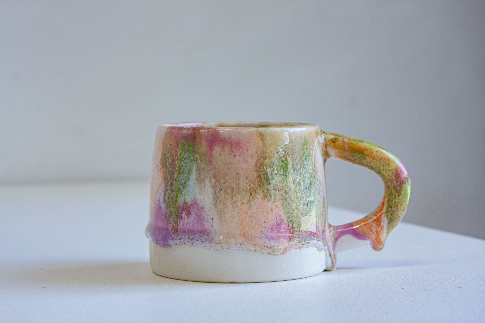 A handmade ceramic mug with a dripping glaze finish, featuring a mix of pink, green, orange, and purple colors on a white base, set against a plain light gray background.
