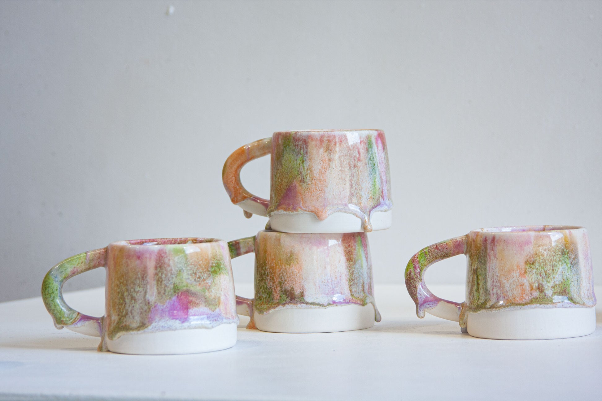 Four handmade ceramic mugs with dripping pastel glaze in pink, green, and purple tones, arranged on a white surface against a light gray background.