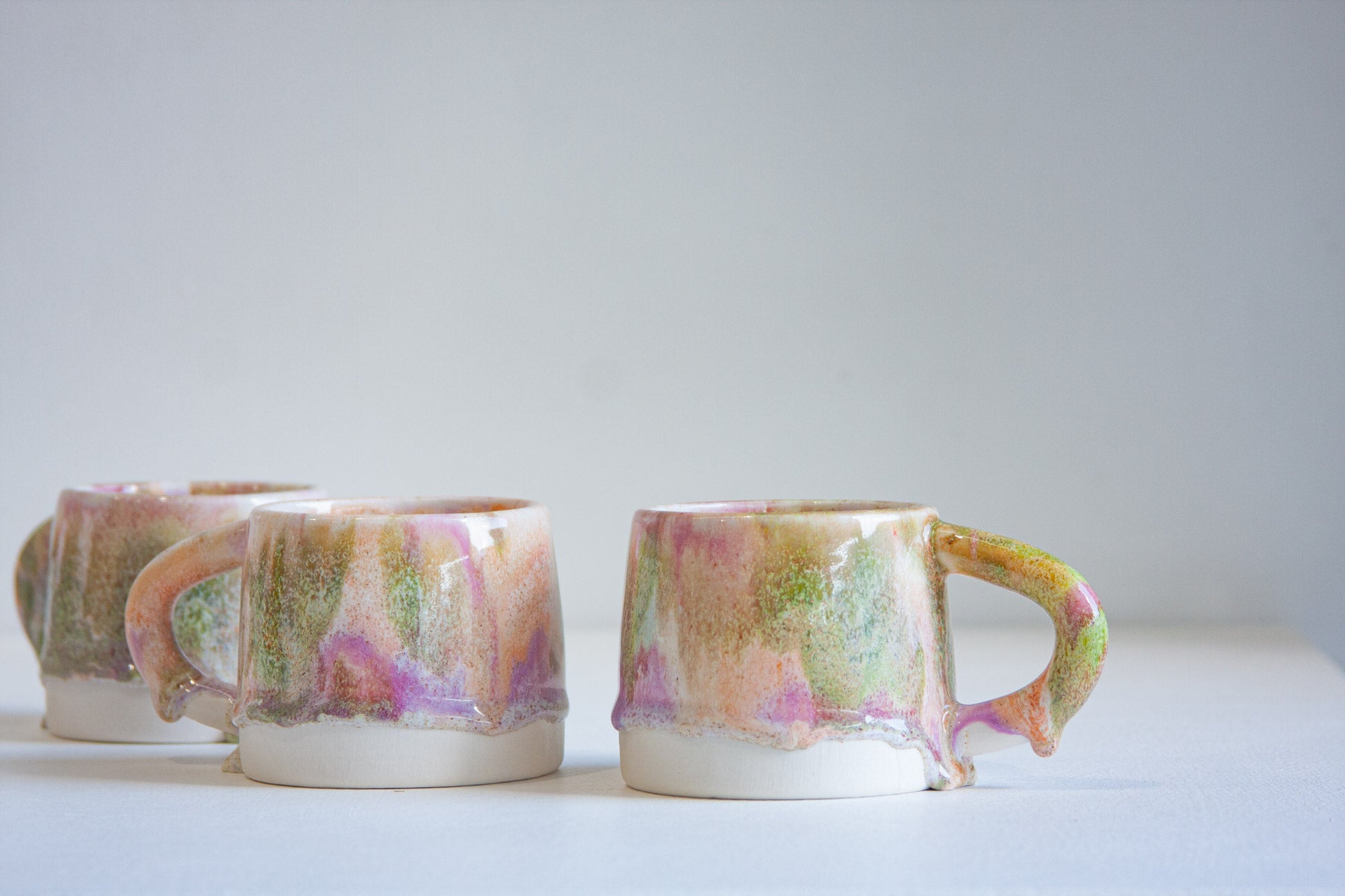 Three handmade ceramic mugs with dripping multicolor glazes in pink, green, and gold tones, arranged on a light gray surface against a neutral gray background.