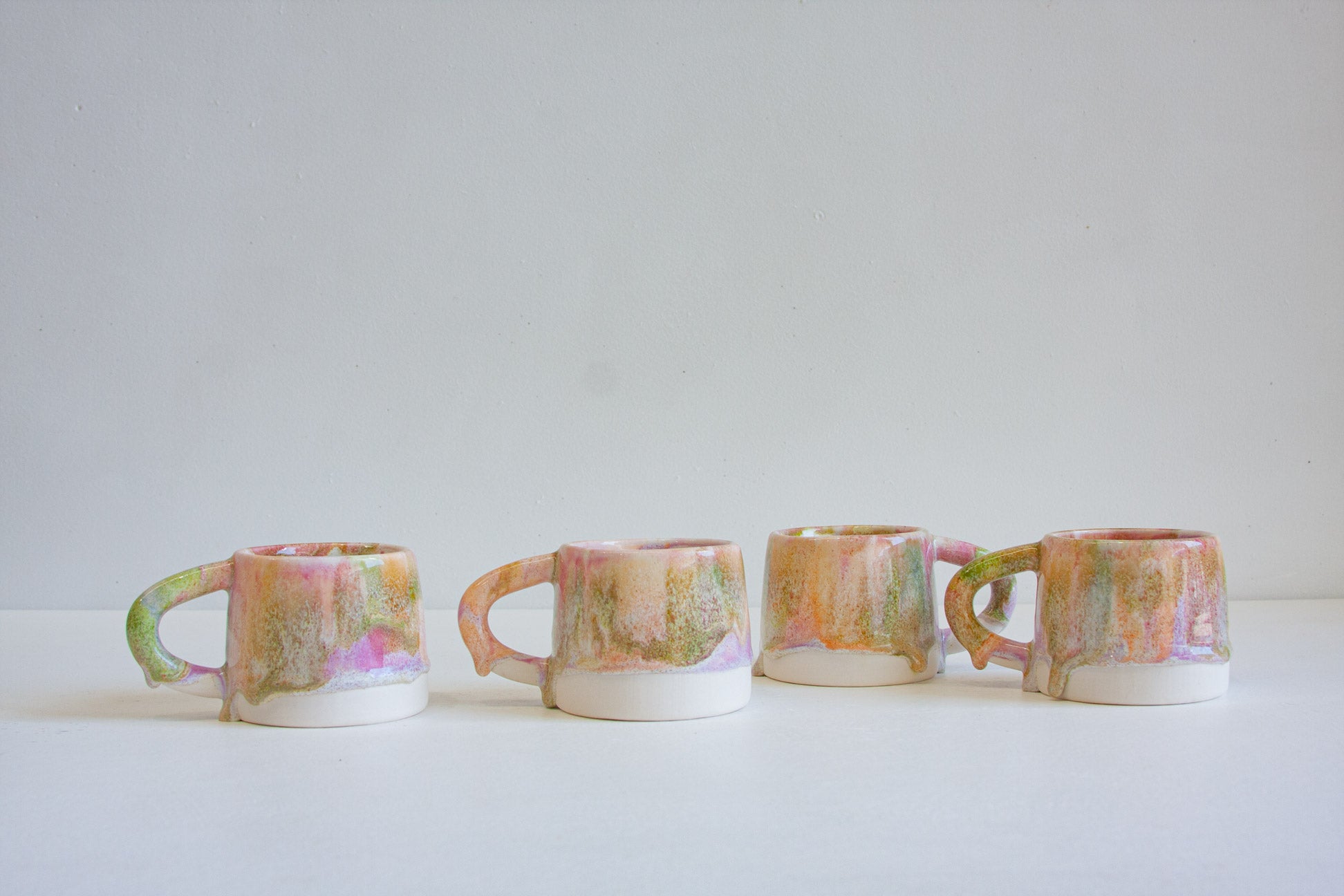 Four small ceramic mugs with handles lined up in a row on a white surface against a light gray background. The mugs feature a marbled, hand-painted glaze with pastel pink, green, orange, and yellow tones over a white base.