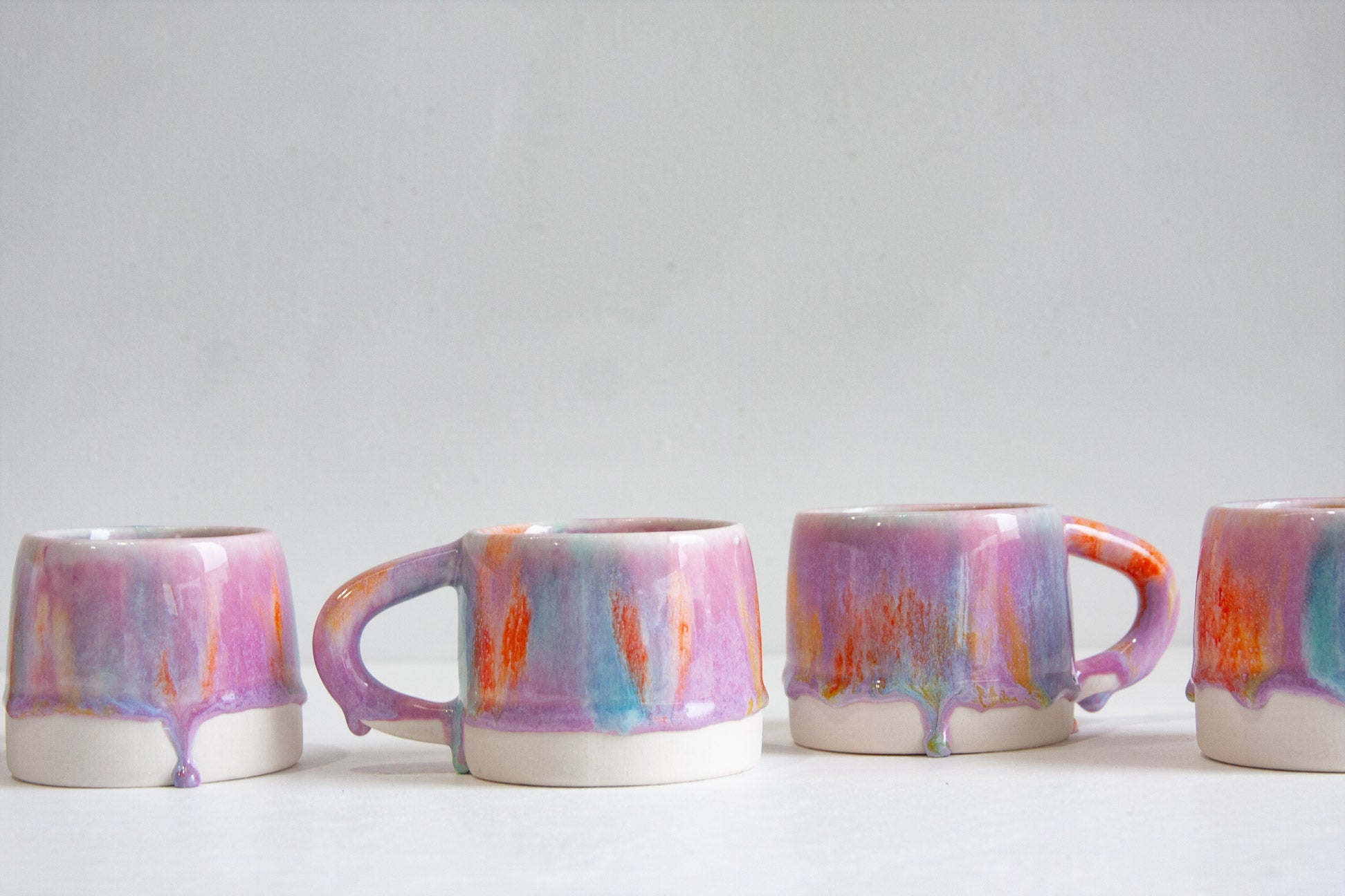 Four handmade ceramic mugs in a row, each glazed in lilac and teal with bright orange drips running down to an unglazed white base.