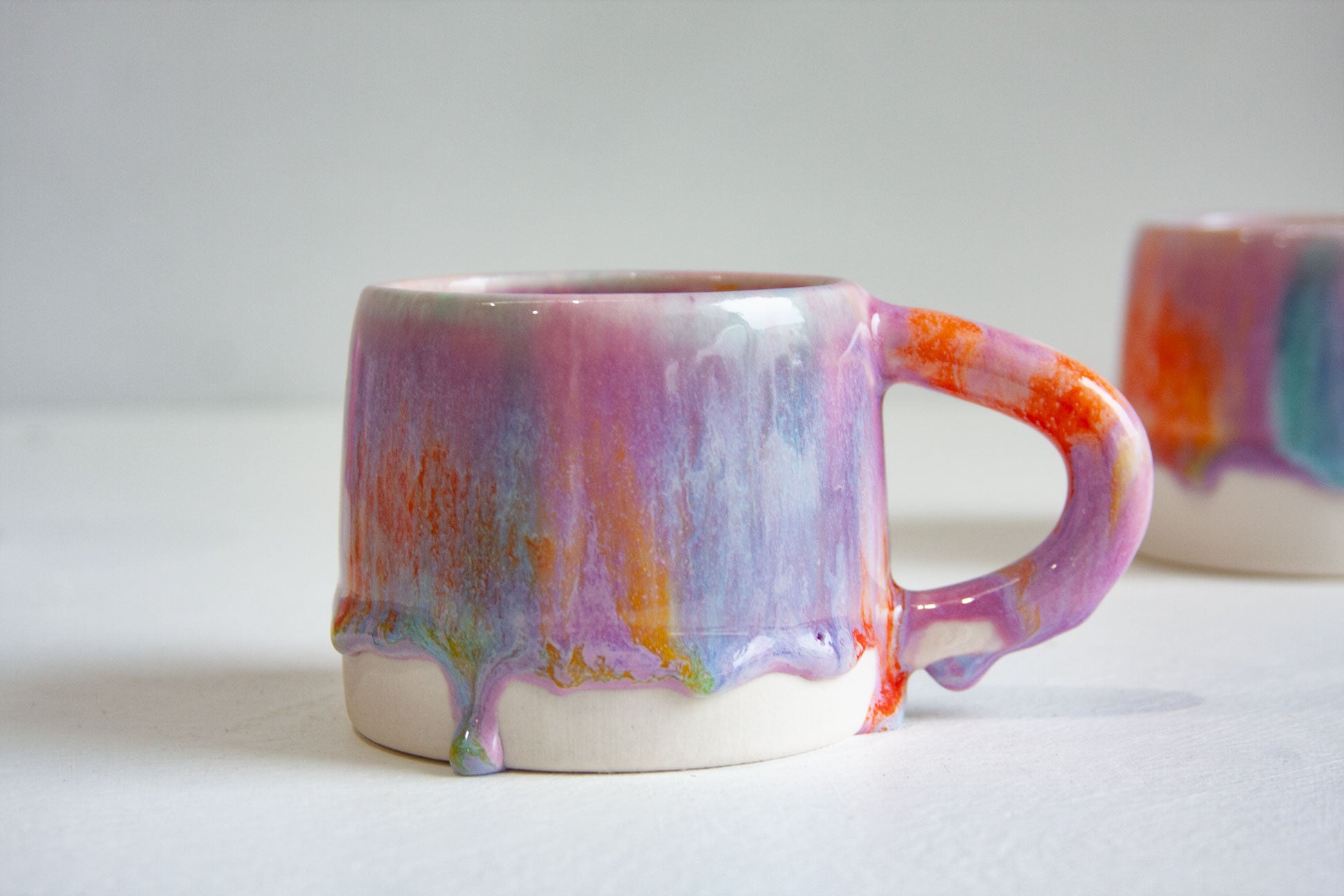 Close-up of a handmade ceramic mug and its handle, lilac and teal glaze streaked with orange, the drips running down and pooling at the unglazed base.