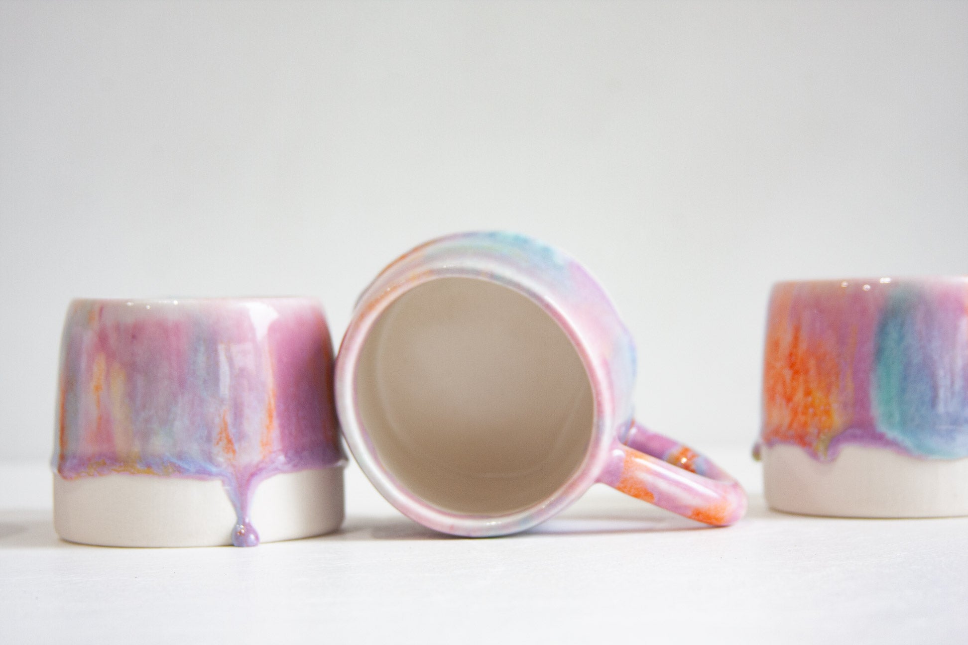 Three handmade ceramic mugs, the middle one on its side showing the plain white glazed interior, the others glazed in pink, lilac, teal and orange.