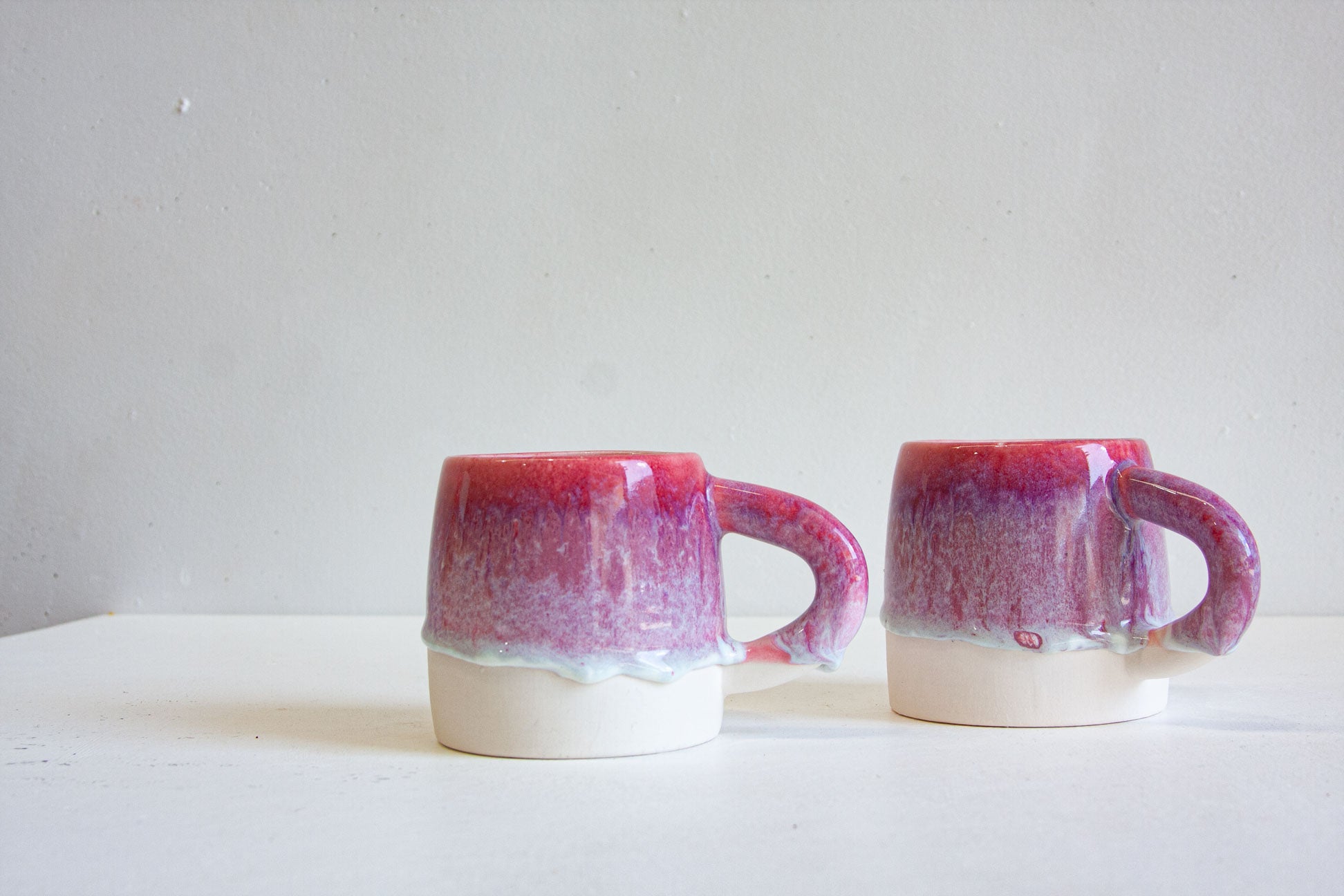 Two handmade ceramic mugs with a glossy dripping glaze in shades of magenta and purple over a white base, set against a plain light gray background.