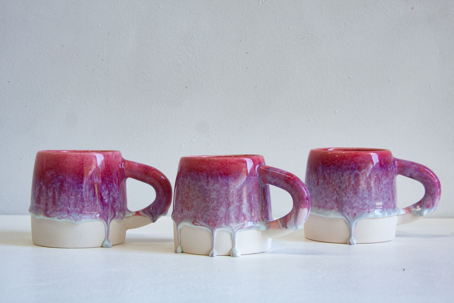 Three ceramic mugs with a glossy dripping glaze finish, featuring a deep magenta and purple top section that fades into a matte off-white base, set against a neutral light gray background.
