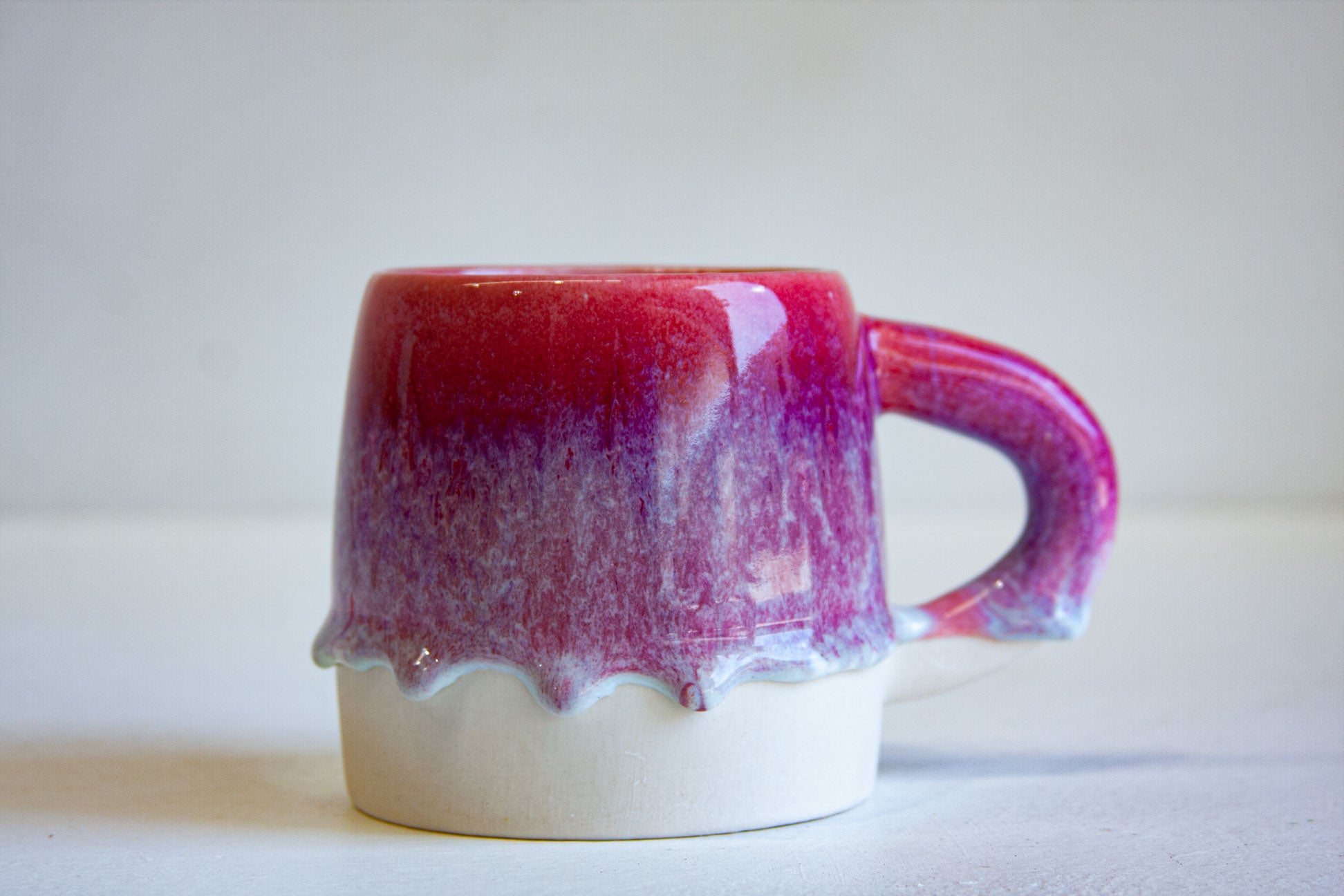 A handmade ceramic mug with a glossy magenta and purple drip glaze on the upper half, a scalloped white clay rim, and a matching sculptural handle, set against a plain light gray background.