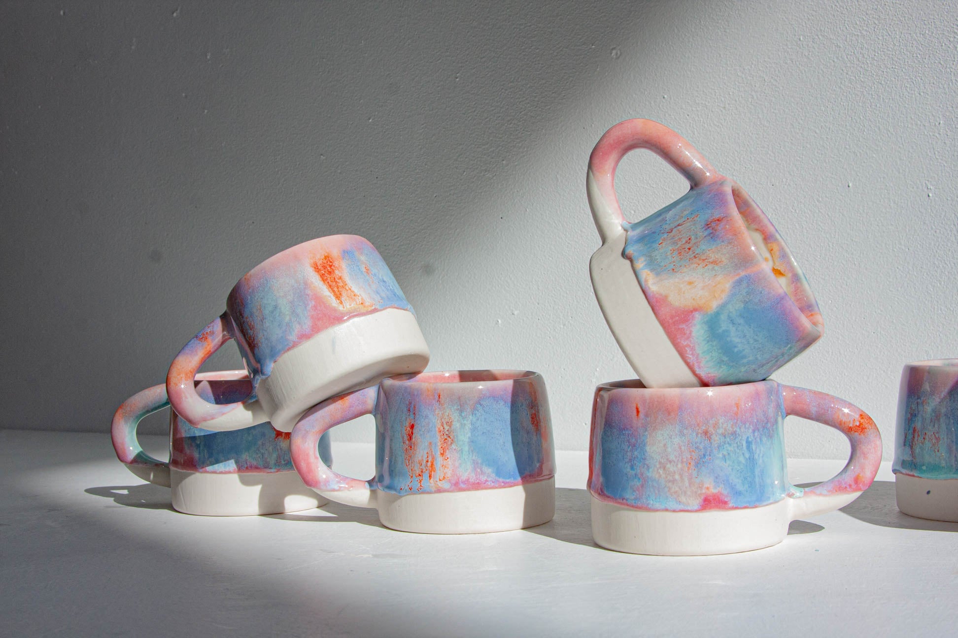 A group of ceramic mugs with a marbled pastel pattern in pink, blue, and orange on a white base, arranged on a white surface against a gray wall.