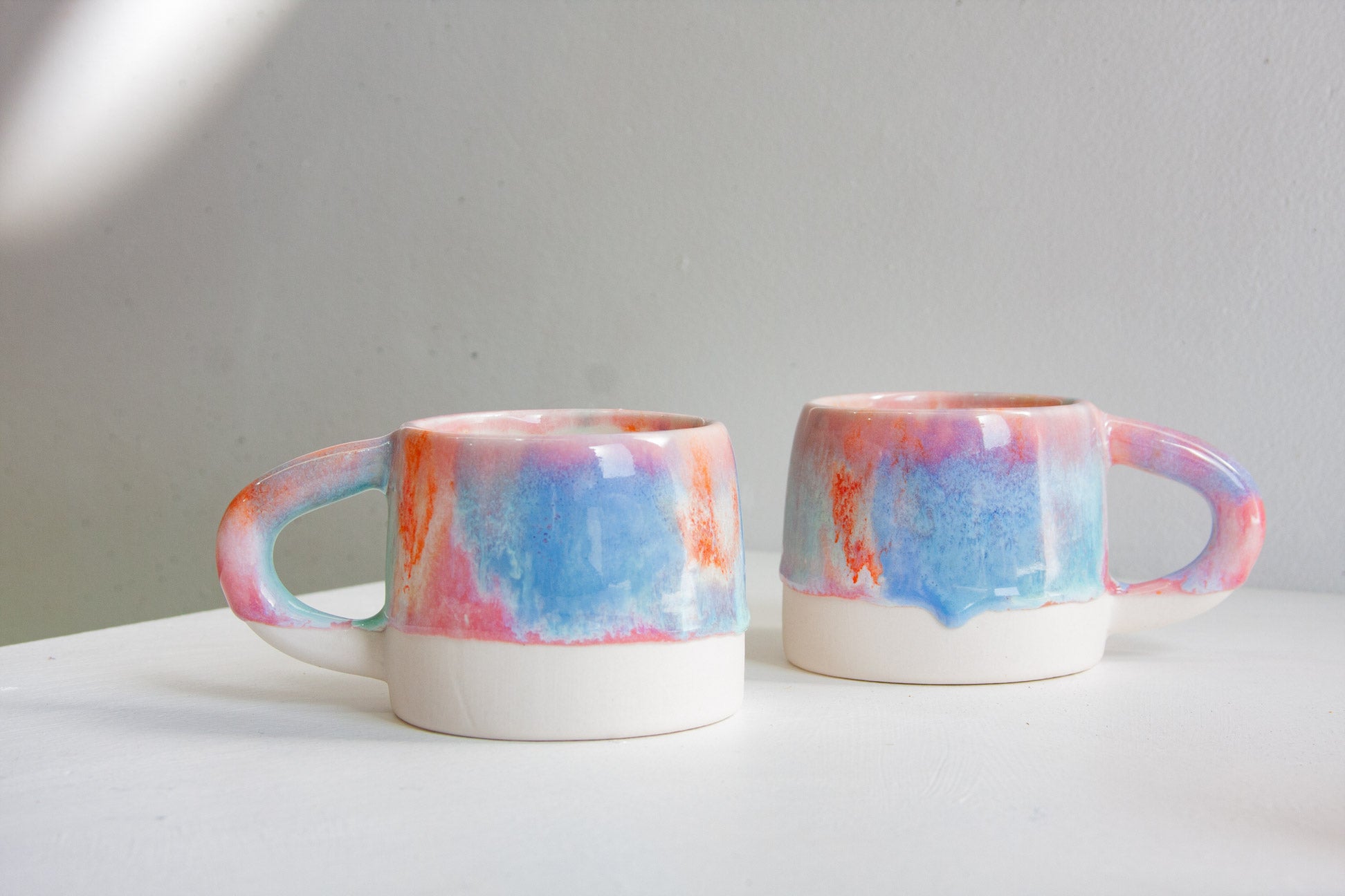 Two ceramic mugs with a glossy white base and a colorful marbled glaze in blue, pink, and orange, displayed on a white surface against a light gray background.