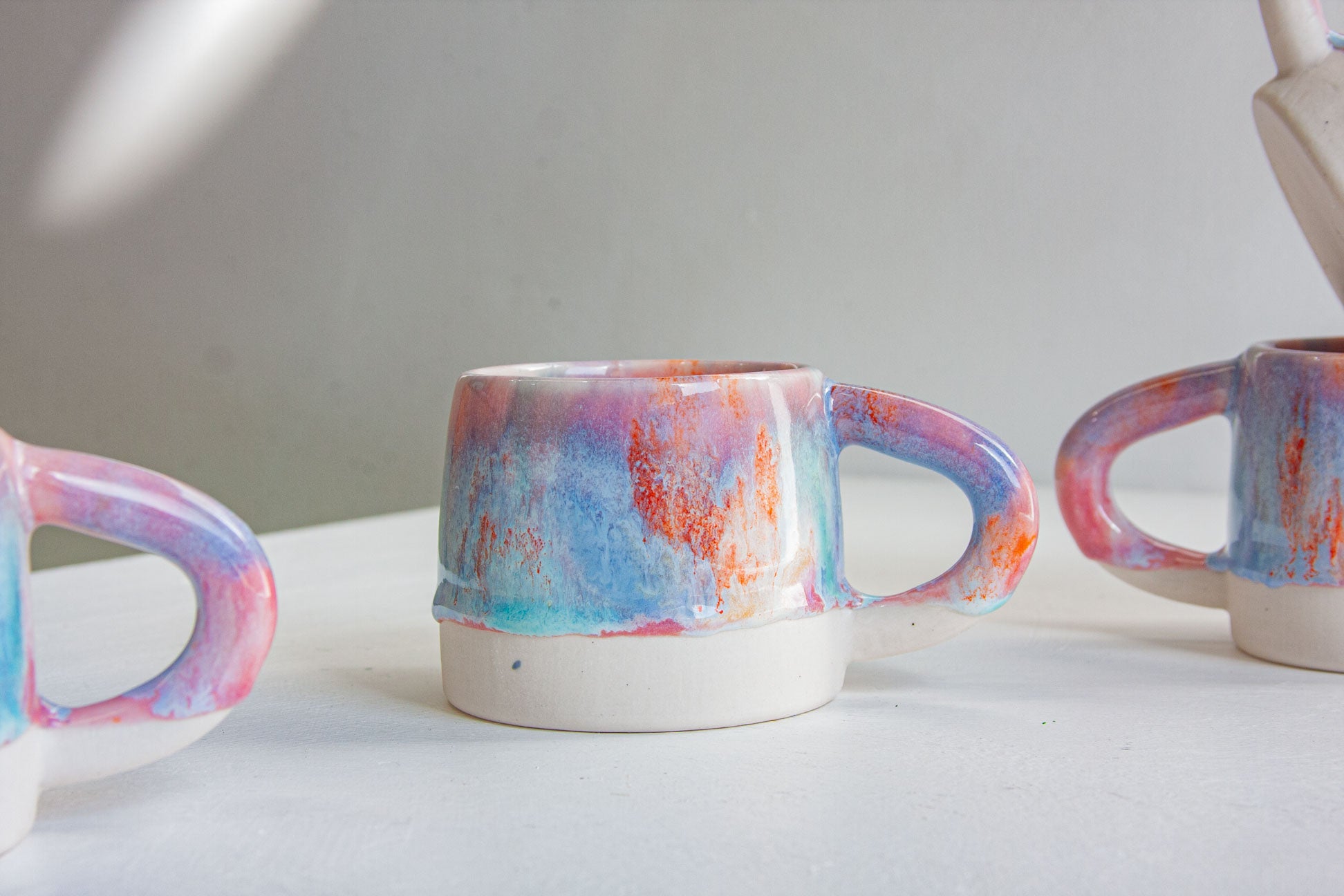 Three handmade ceramic mugs with a colorful marbled glaze in blue, pink, purple, and orange, displayed on a light gray surface.