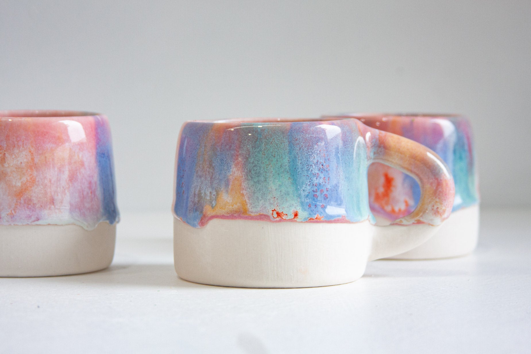 Three handmade ceramic mugs with a glossy multicolor drip glaze in shades of blue, pink, purple, and orange, paired with a matte off-white base, arranged on a light gray surface.