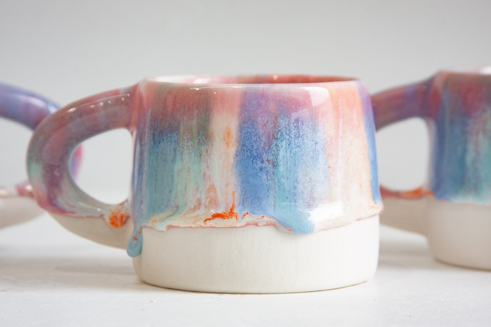 A ceramic mug with a dripping glaze design, featuring a white base and a colorful upper section with blue, pink, purple, and orange tones, shown on a light gray background.