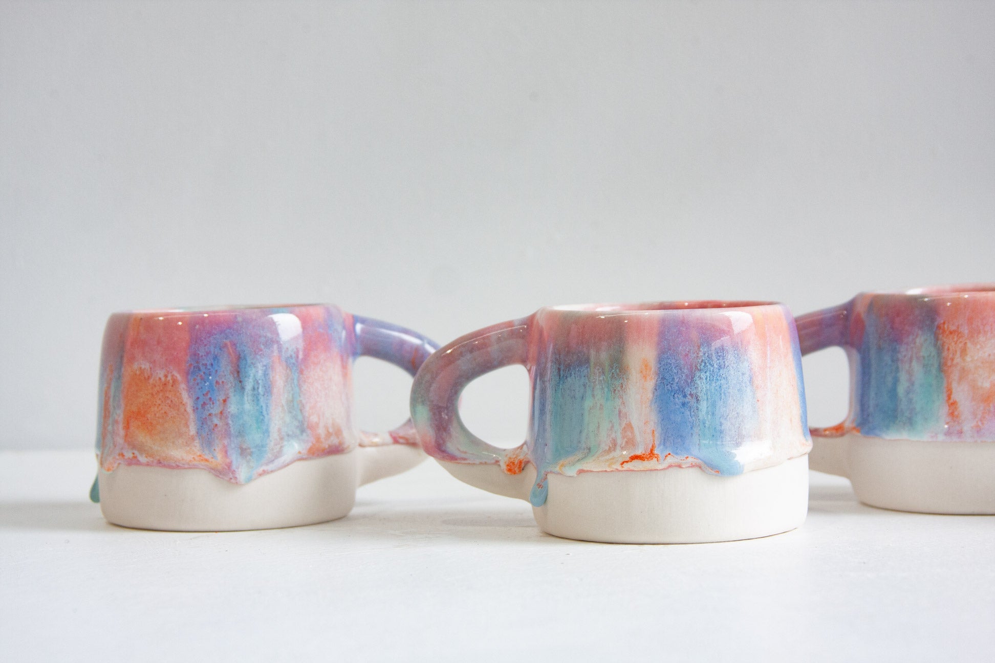 Three ceramic mugs with dripping multicolor glaze in shades of blue, pink, purple, and orange, set against a plain light gray background.