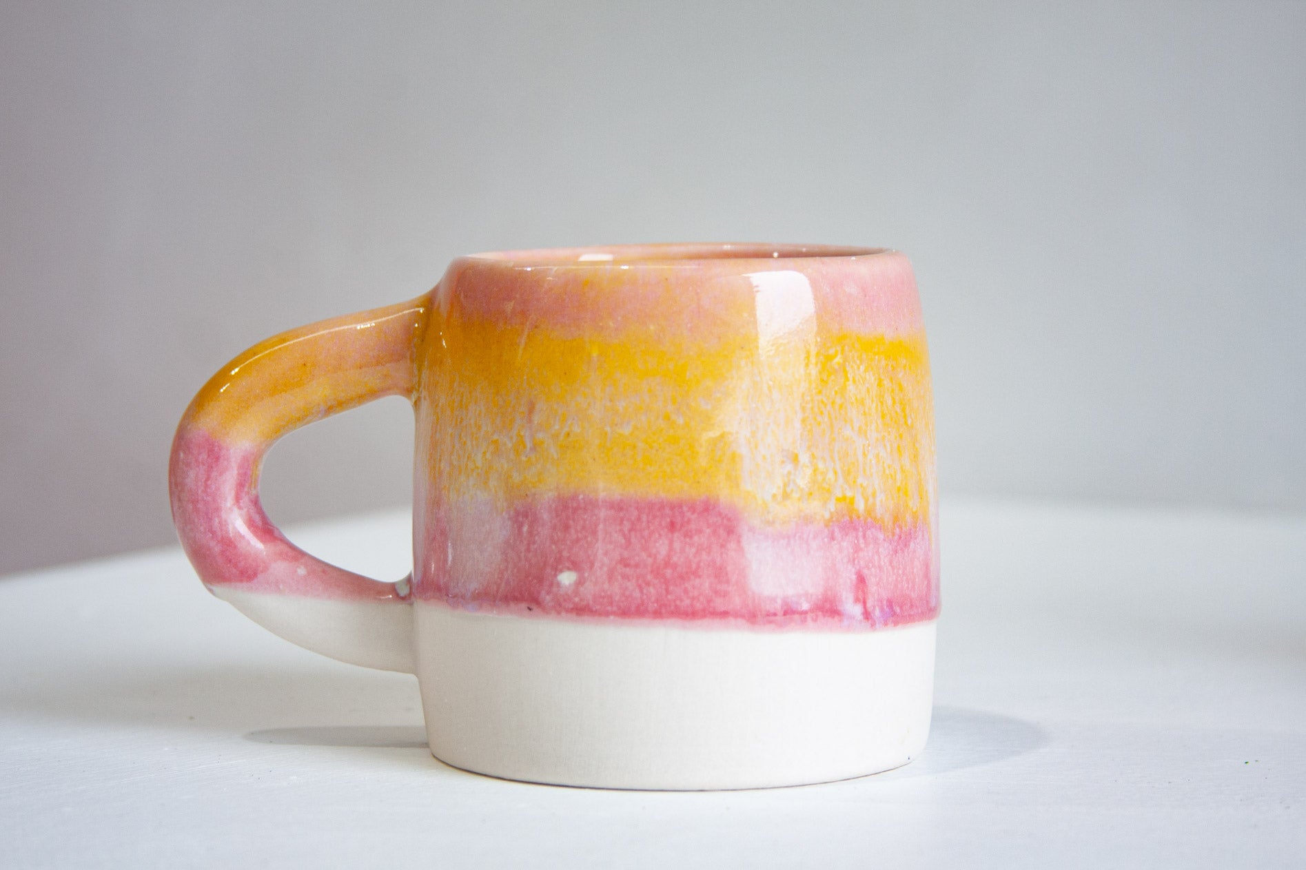 A handmade ceramic large mug seen from the side, pink at the rim over a band of yellow and a band of pink, meeting an unglazed white base.