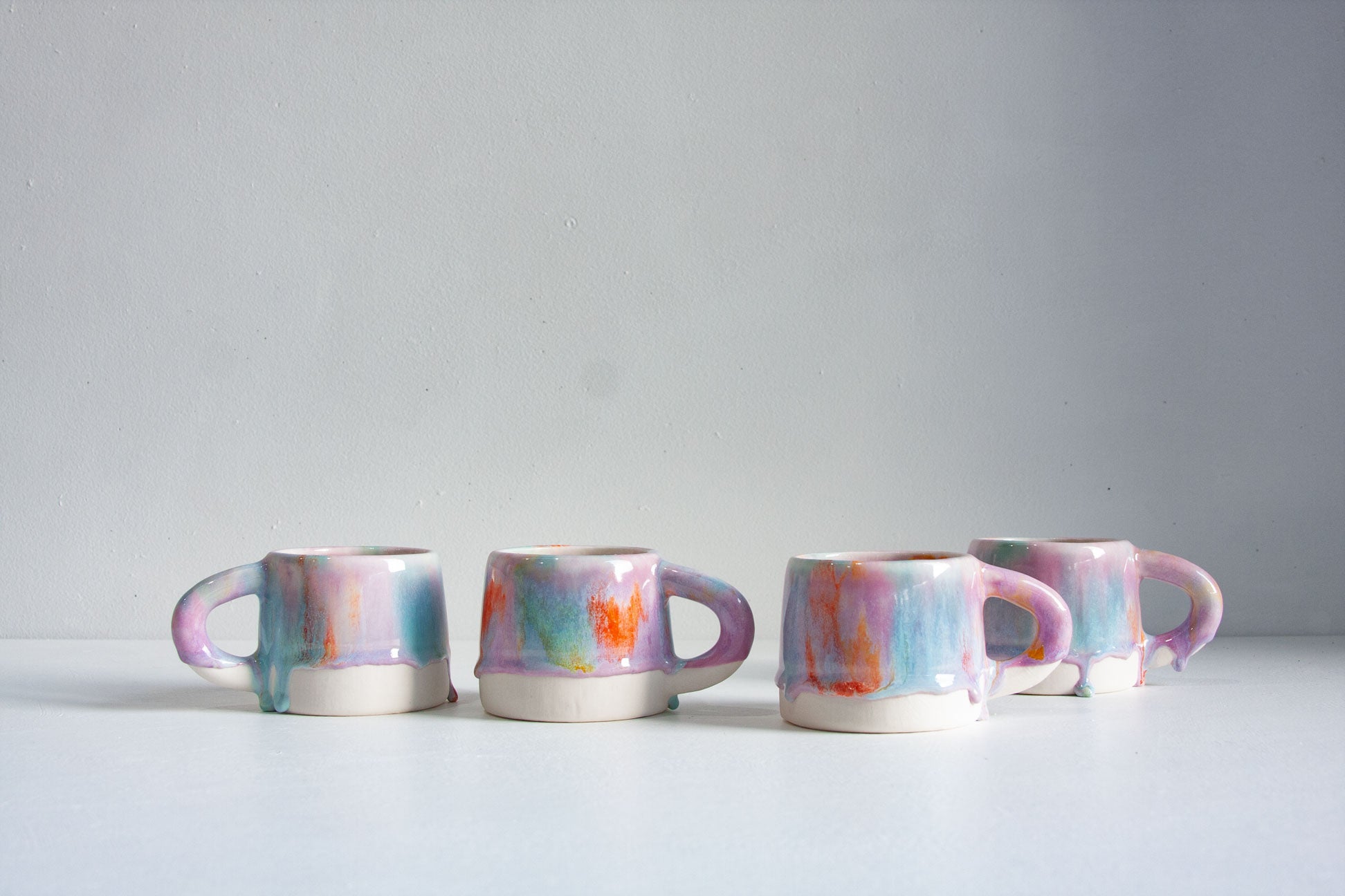 Four small ceramic mugs with colourful dripping glaze patterns in pink, blue, purple, and orange, arranged on a white surface against a light grey background.