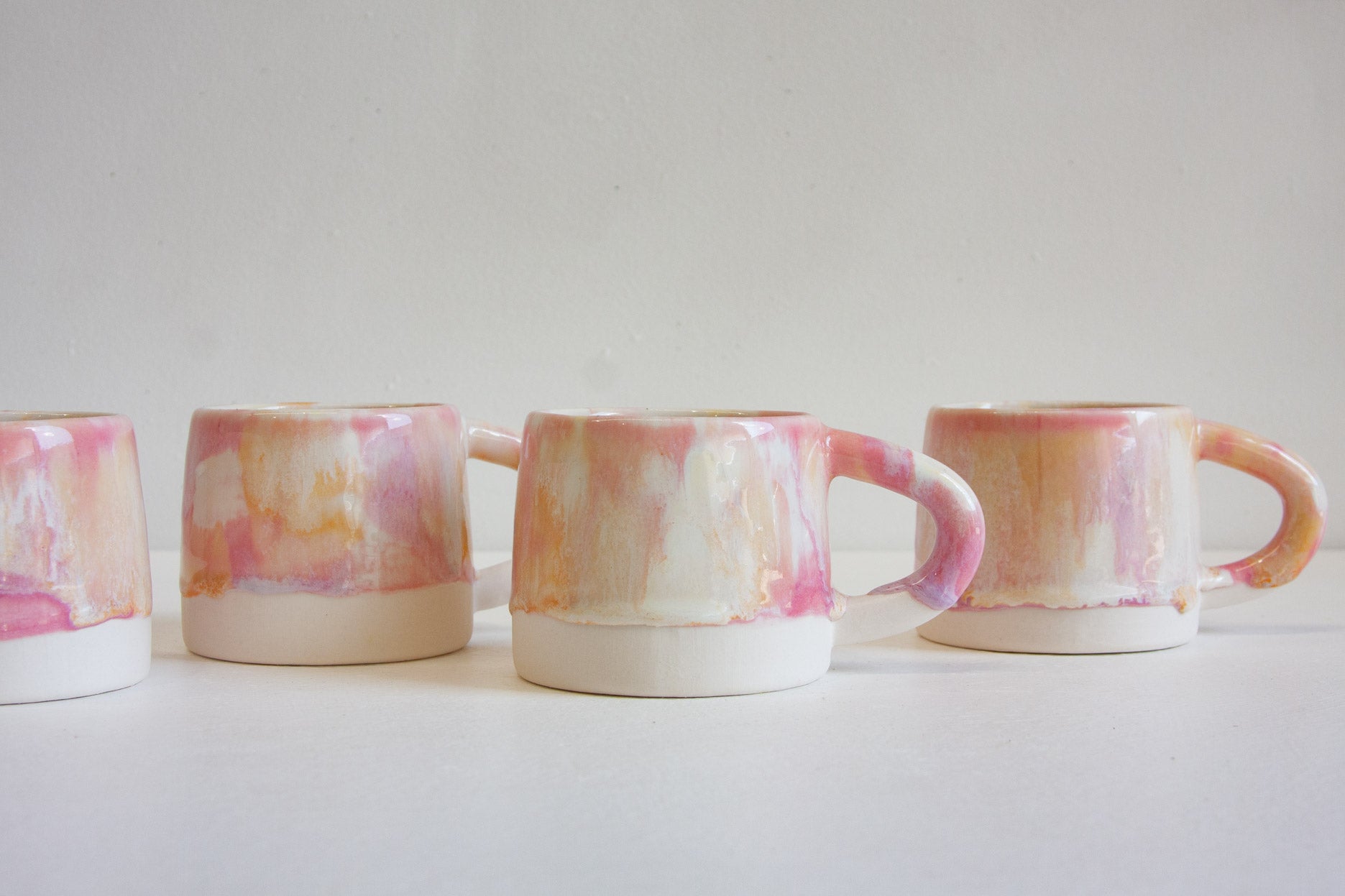 Four handmade ceramic mugs with handles, featuring a marbled pink, peach, and white glaze, lined up on a white surface against a light gray background.
