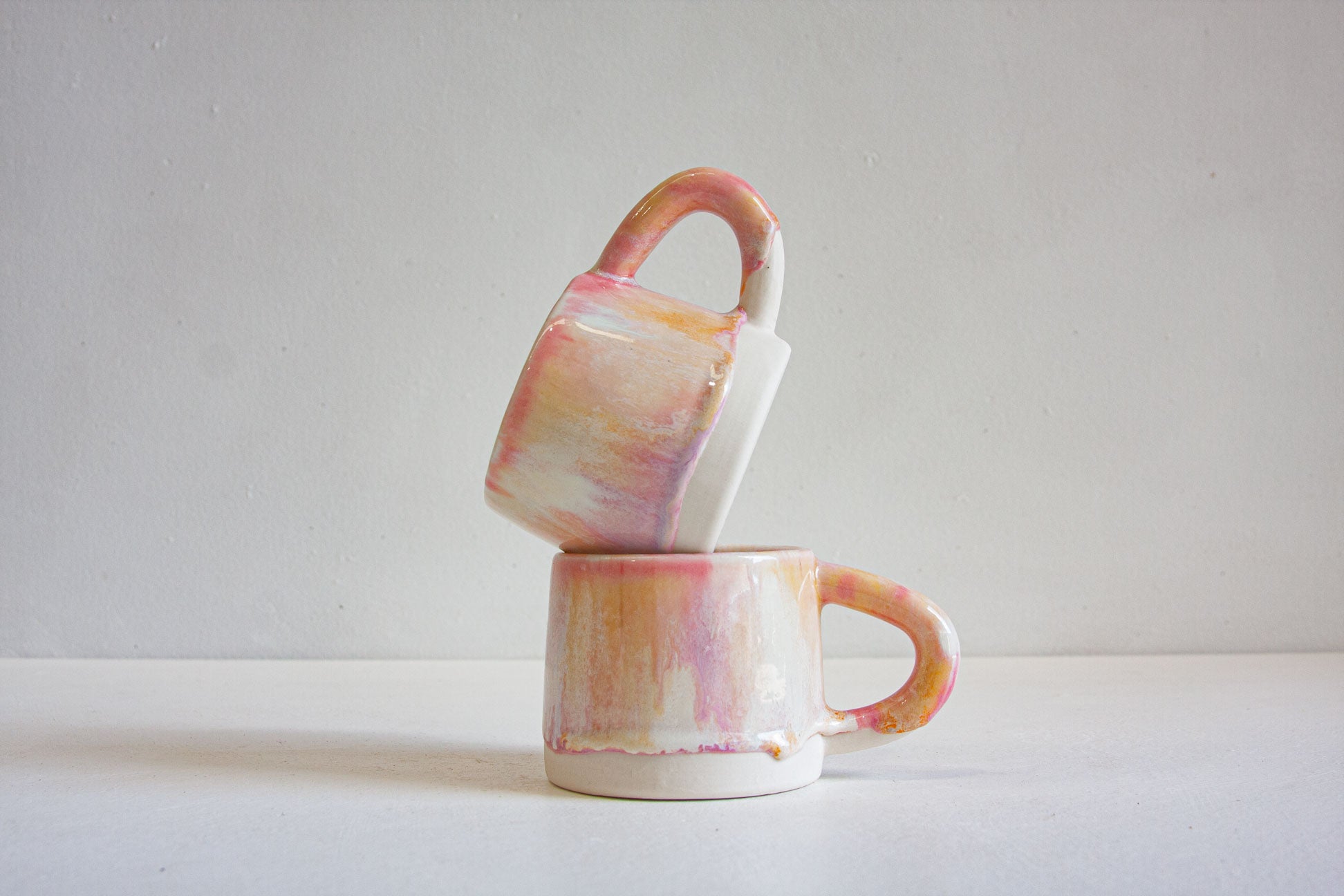 Two small ceramic mugs with abstract pink, peach, and cream glaze stacked on a white surface against a light gray background.