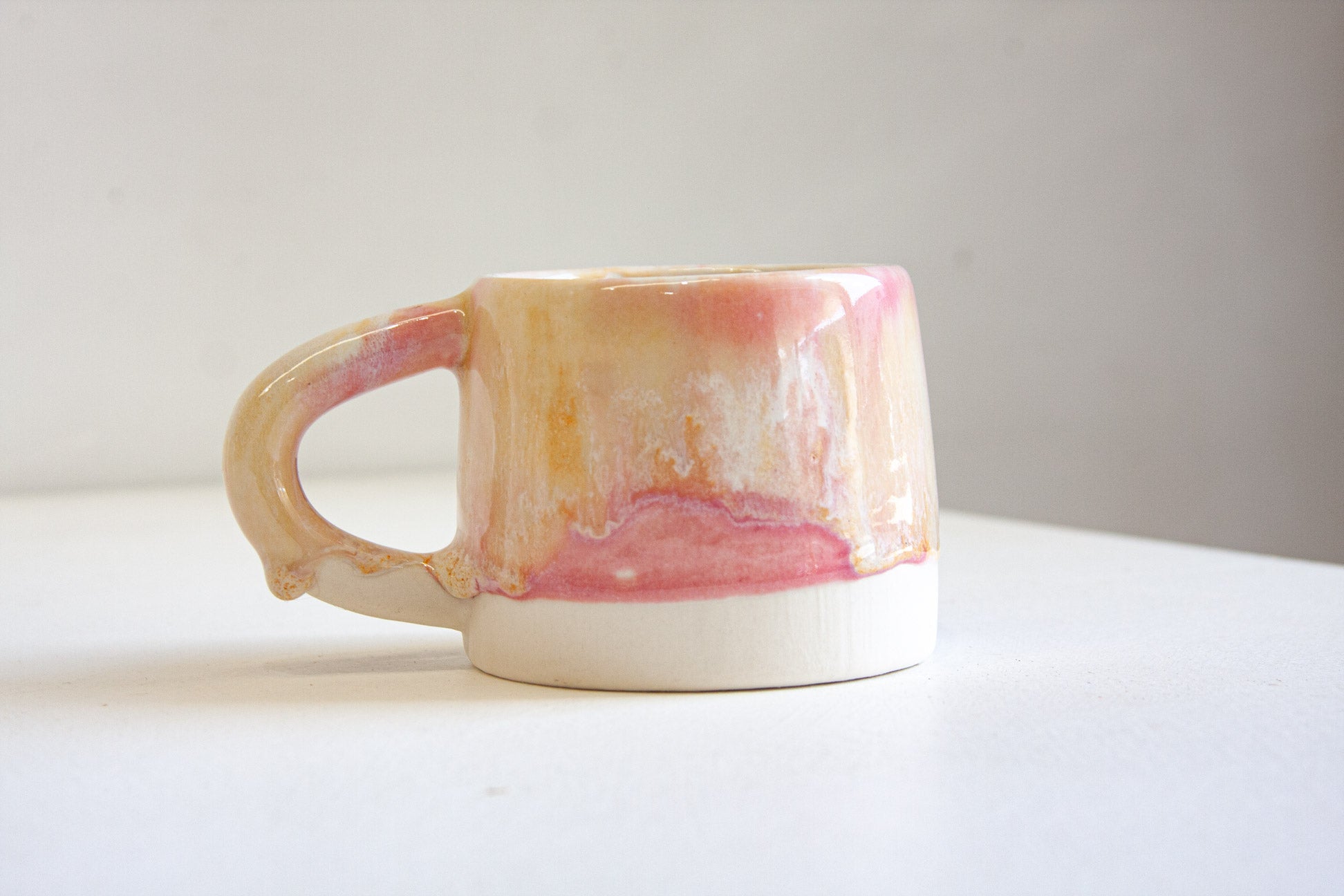 A handmade ceramic mug with a glossy finish, featuring a marbled pattern of pink, orange, and cream glaze over a white base, set against a plain light gray background.