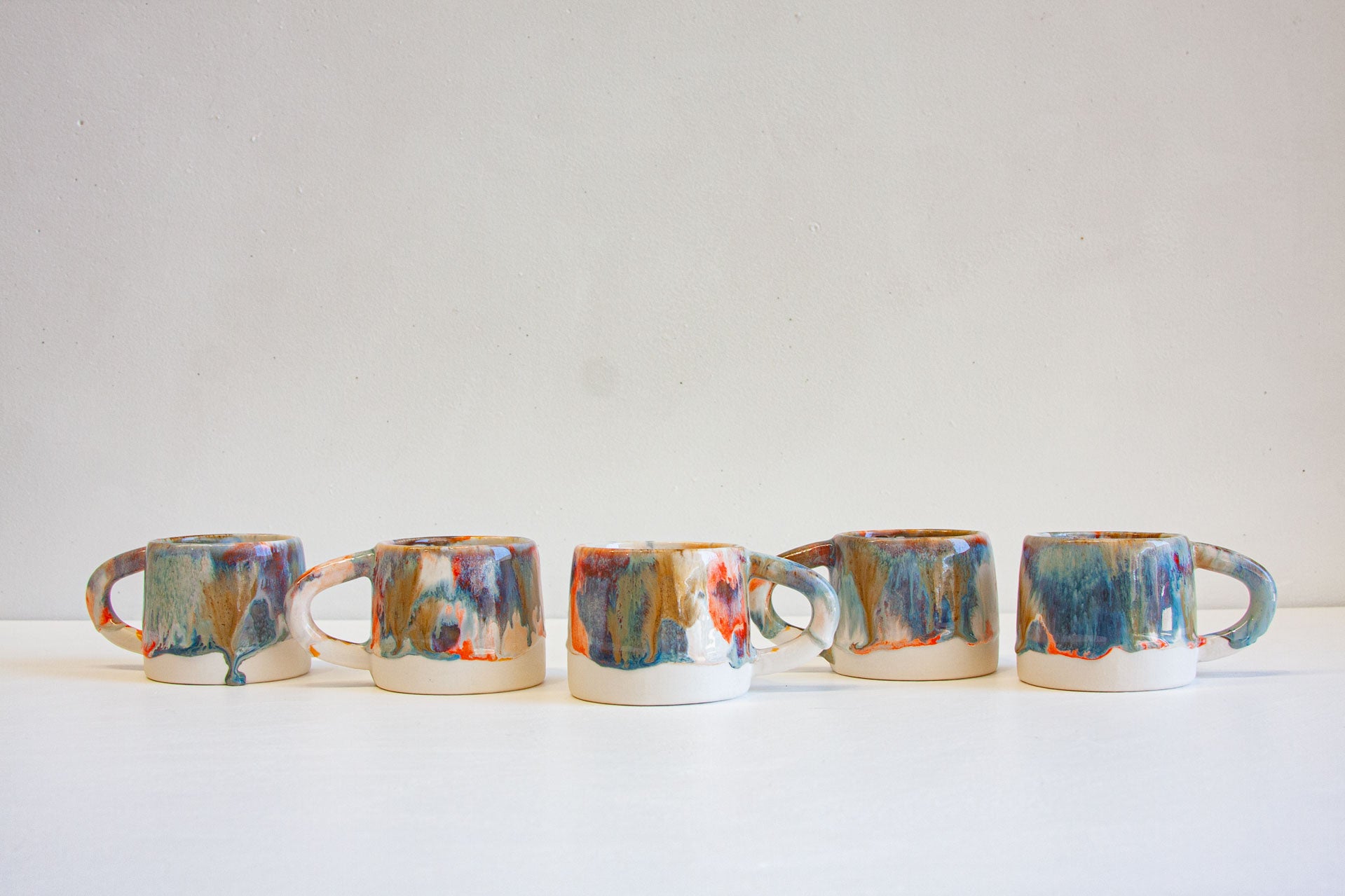 Five ceramic mugs with colorful abstract dripping glazes in blue, orange, red, and green on a white base, arranged in a row against a plain off-white background.
