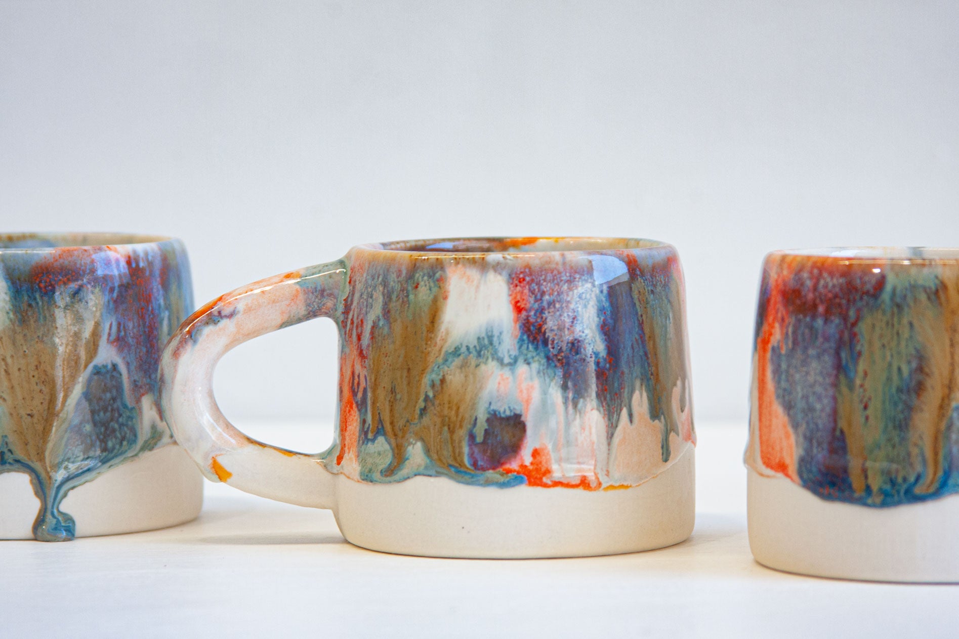 Three ceramic mugs with a colorful dripping glaze pattern in blue, orange, red, and brown on a white base, photographed against a clean white background.