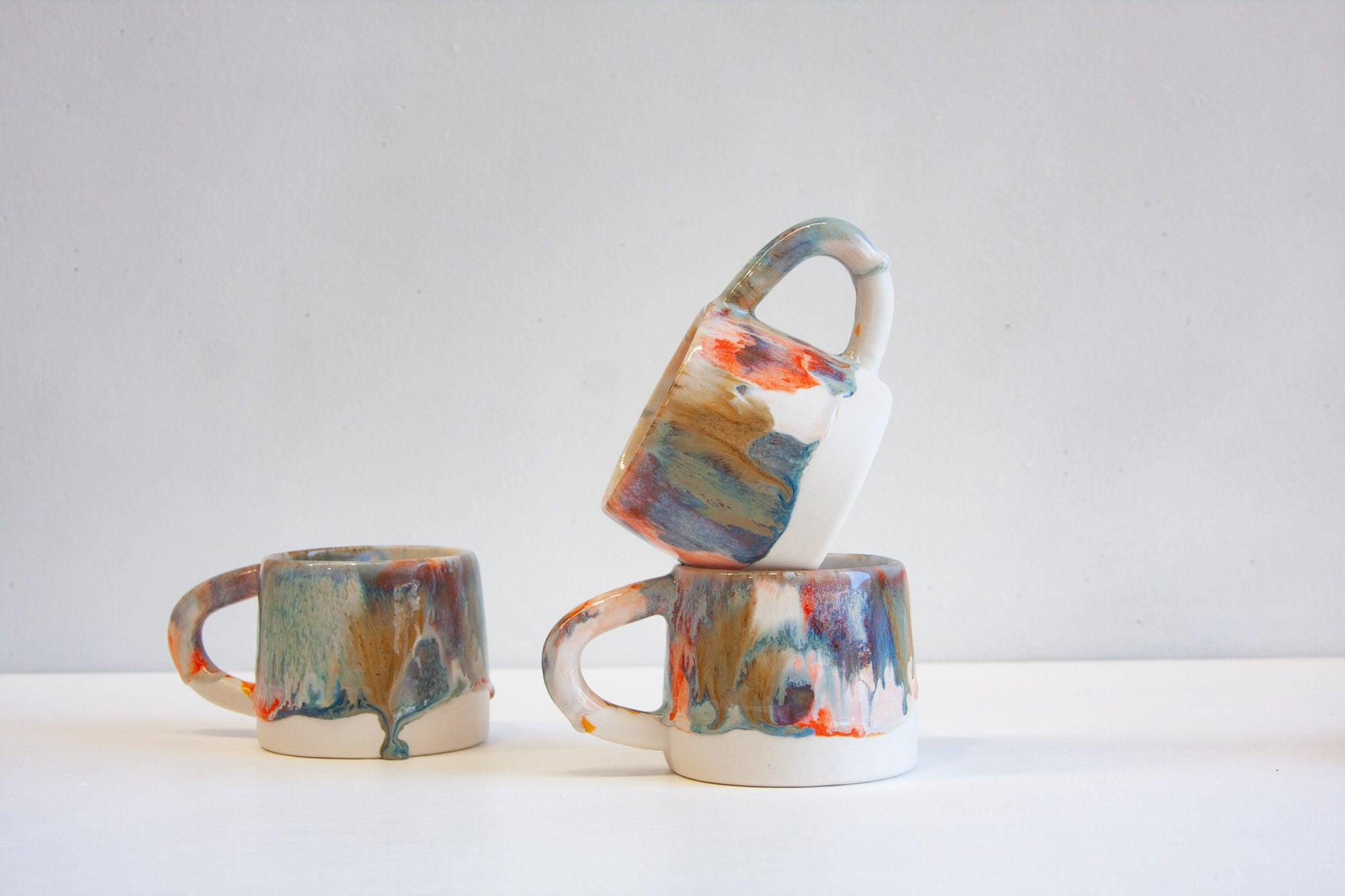 Three small ceramic mugs with abstract marbled paint patterns in blue, orange, brown, and white, arranged on a white surface against a light gray background.