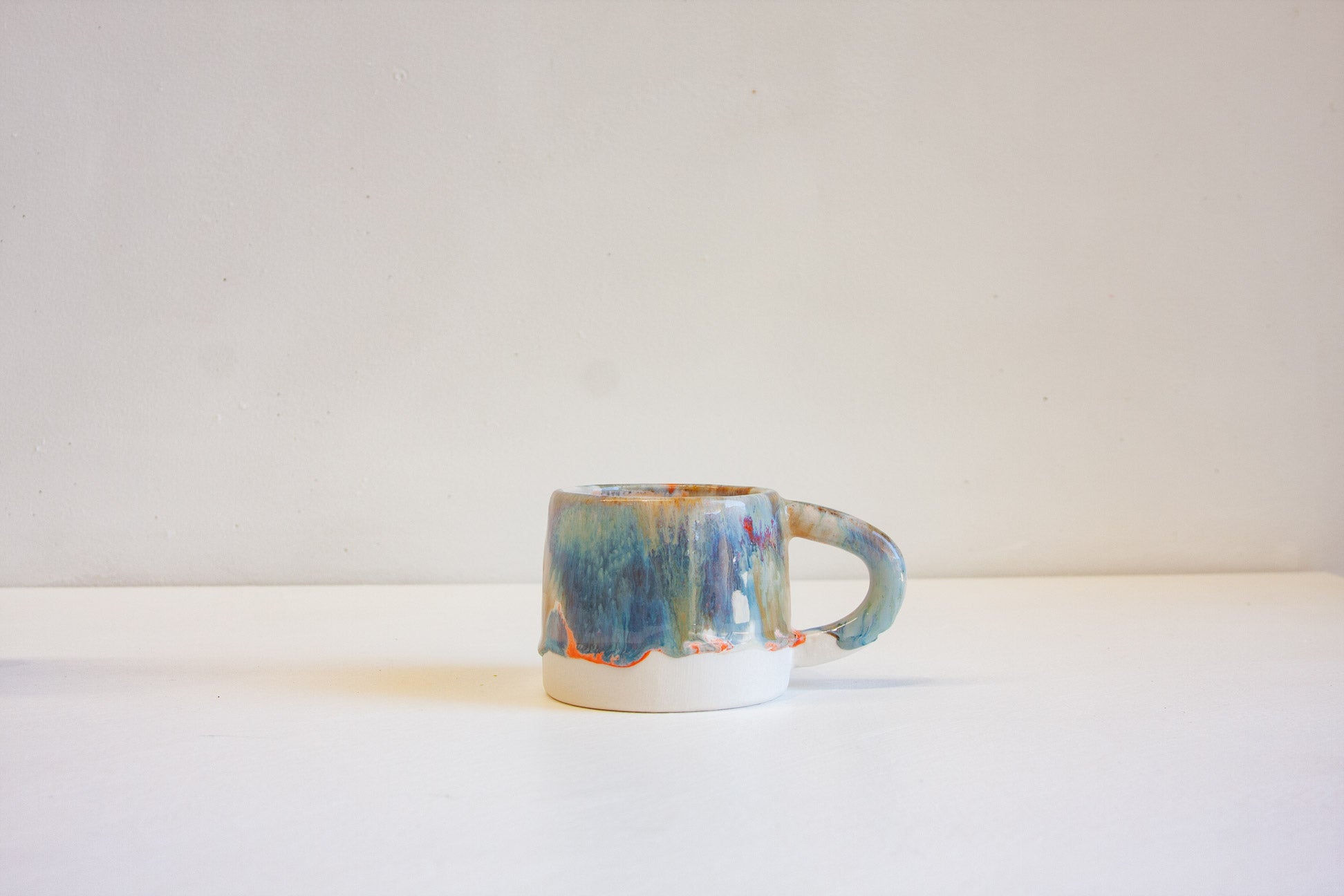 A small ceramic mug with a handle, featuring a textured, multicolored glaze in shades of blue, green, and orange, sitting on a white surface against a plain off-white background.