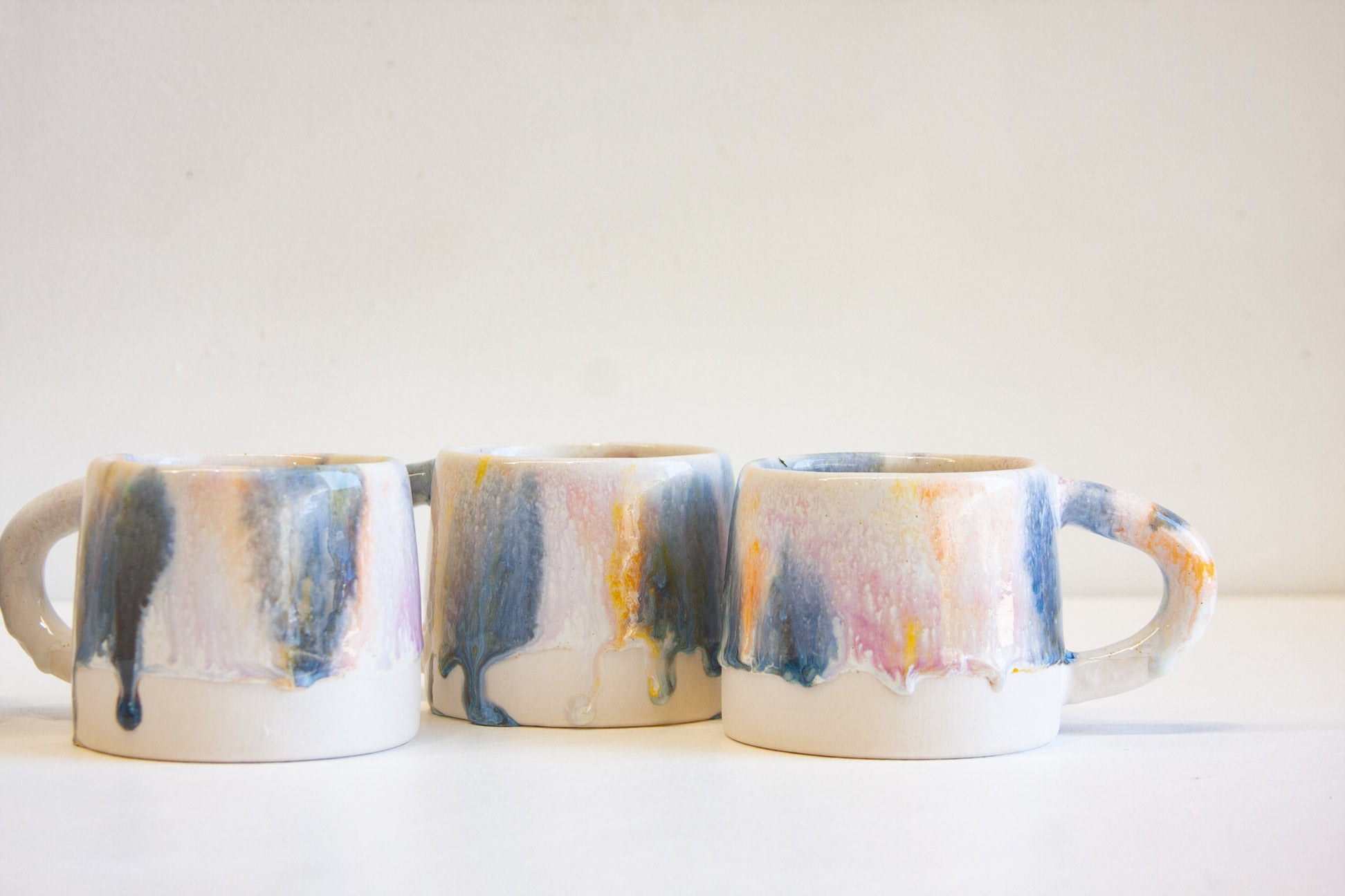 Three ceramic mugs with dripping multicolor glazes in blue, pink, yellow, and gray on a white base, arranged side-by-side against a plain off-white background.