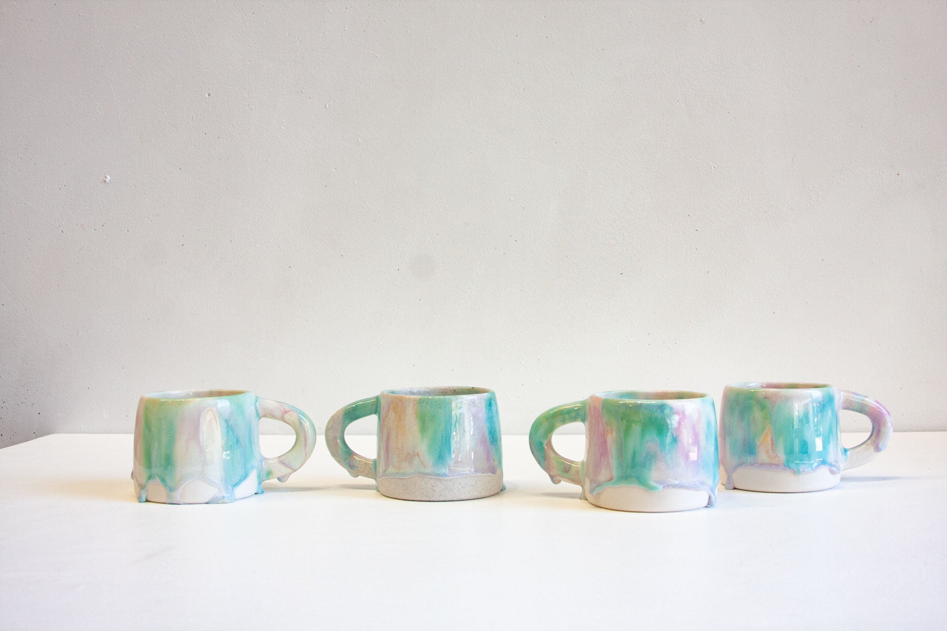 Four small ceramic mugs with handles, decorated with a marbled pastel glaze of turquoise, mint green, lavender, and white, lined up on a white surface against a light gray wall.