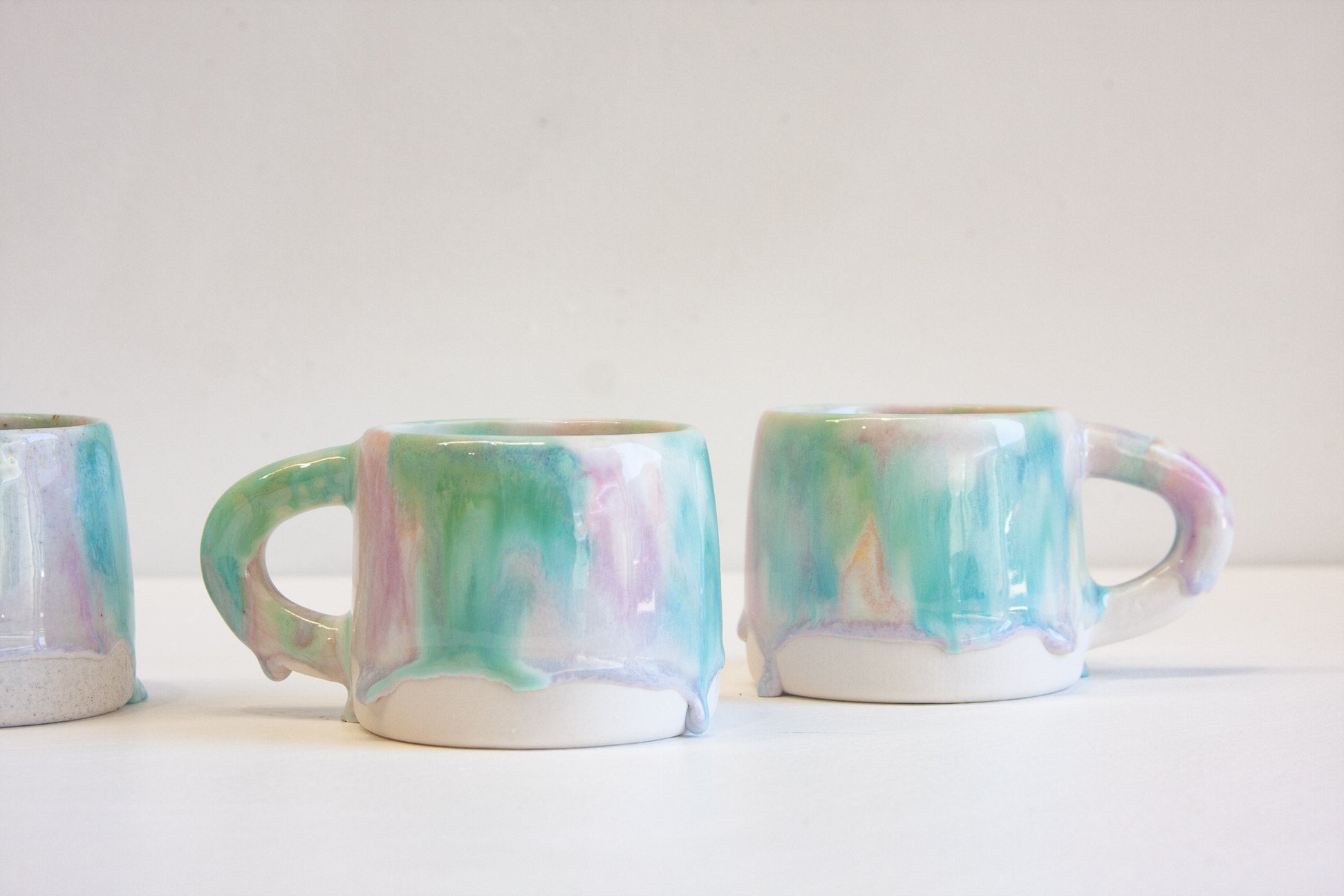 Three handmade ceramic mugs with dripping pastel glazes in mint green, pink, and lavender on a white surface against a light gray background.