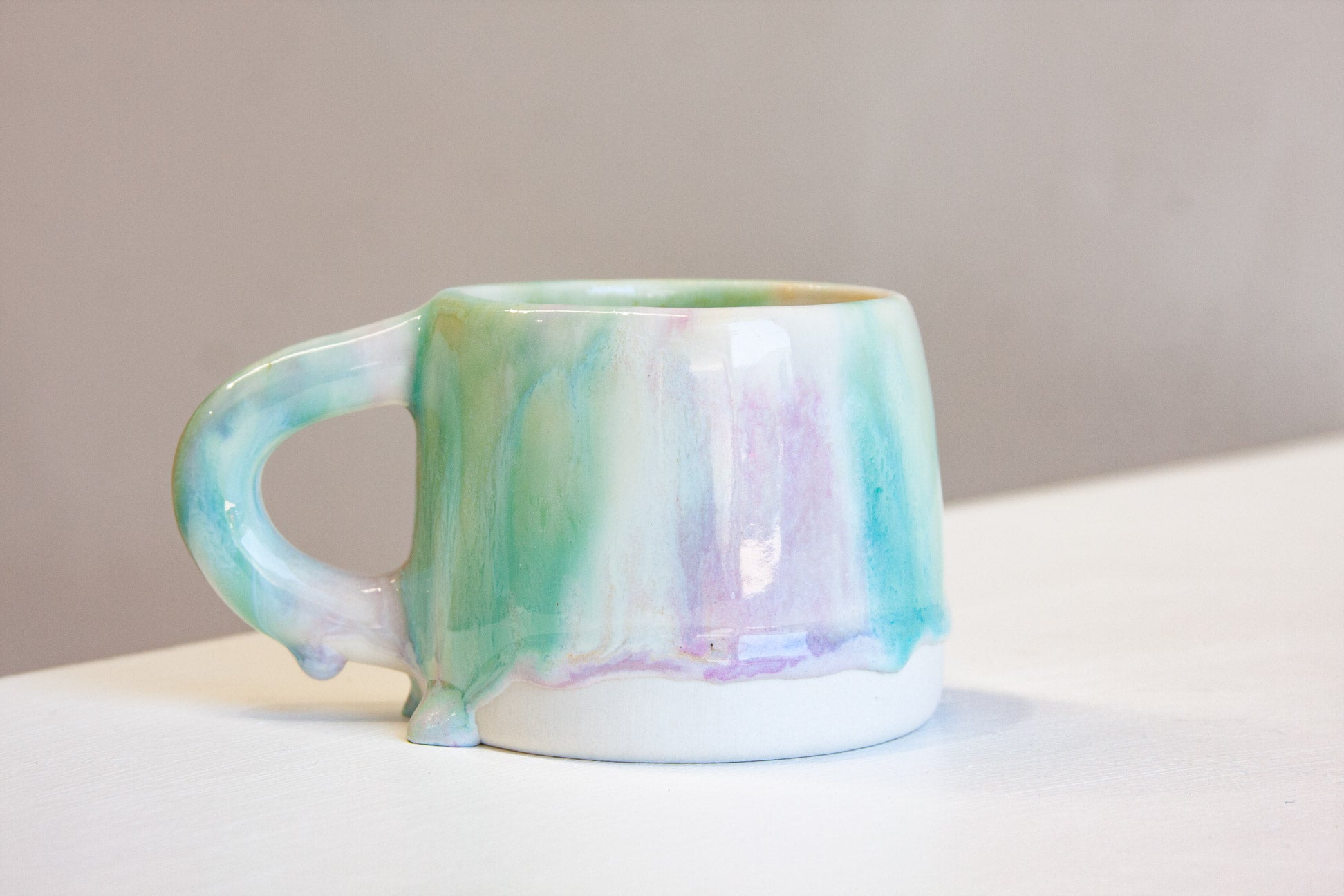 A ceramic mug with a dripping glaze finish, featuring pastel shades of mint green, lavender purple, and soft blue on a white base, set against a neutral gray background.