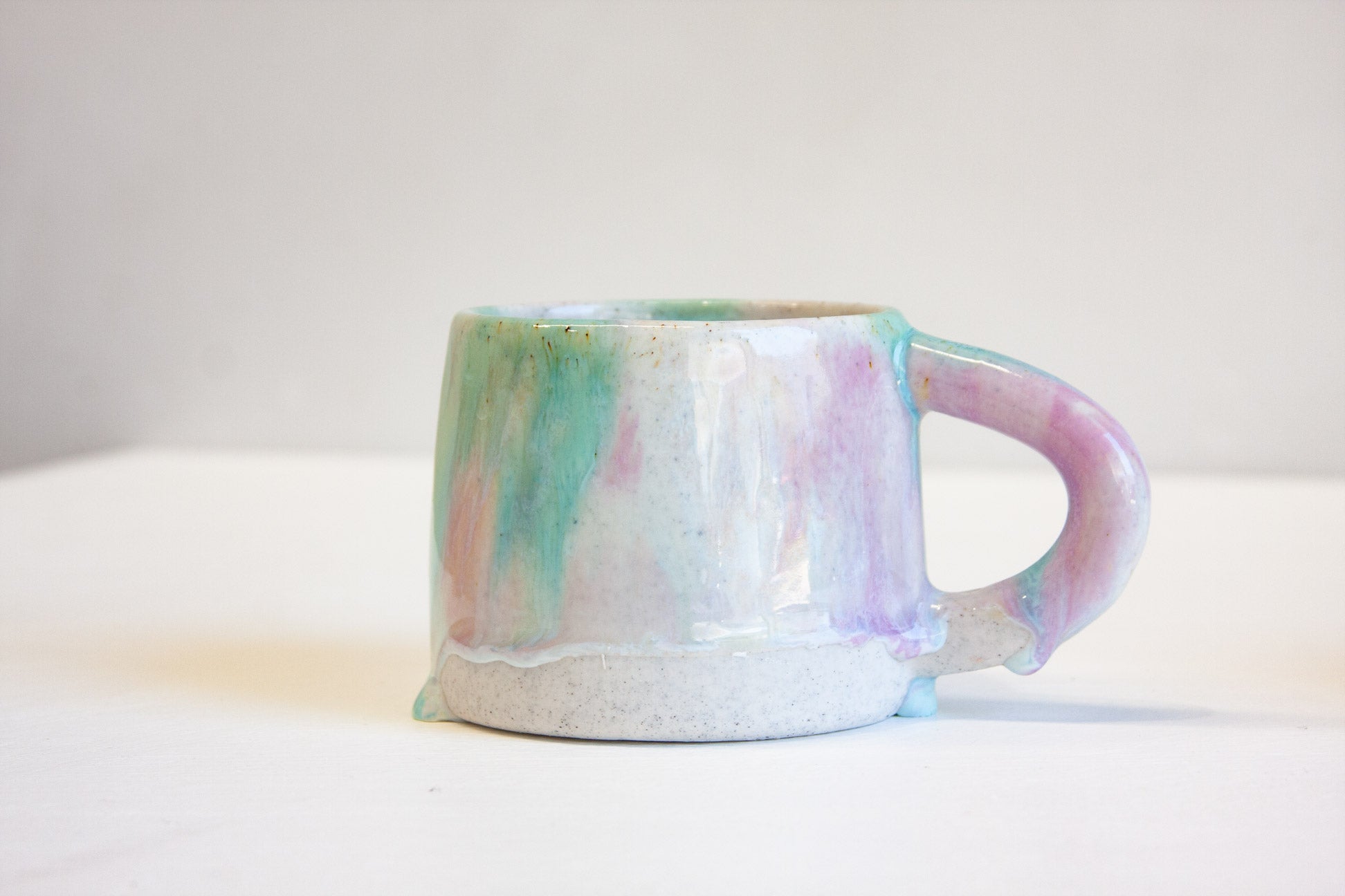 A handmade ceramic mug with a pastel marbled glaze featuring pink, green, and blue tones on a white base, set against a plain light gray background.