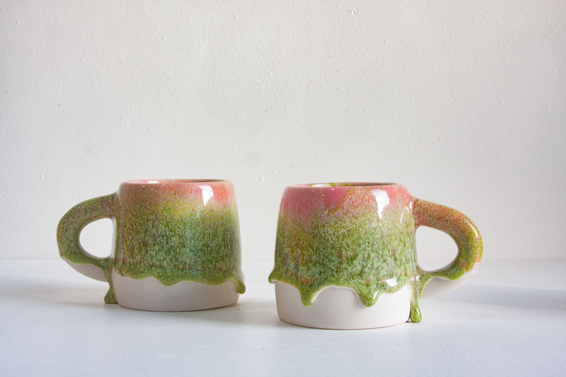 Two handmade ceramic pink and green drippy glaze mugs on a white table.