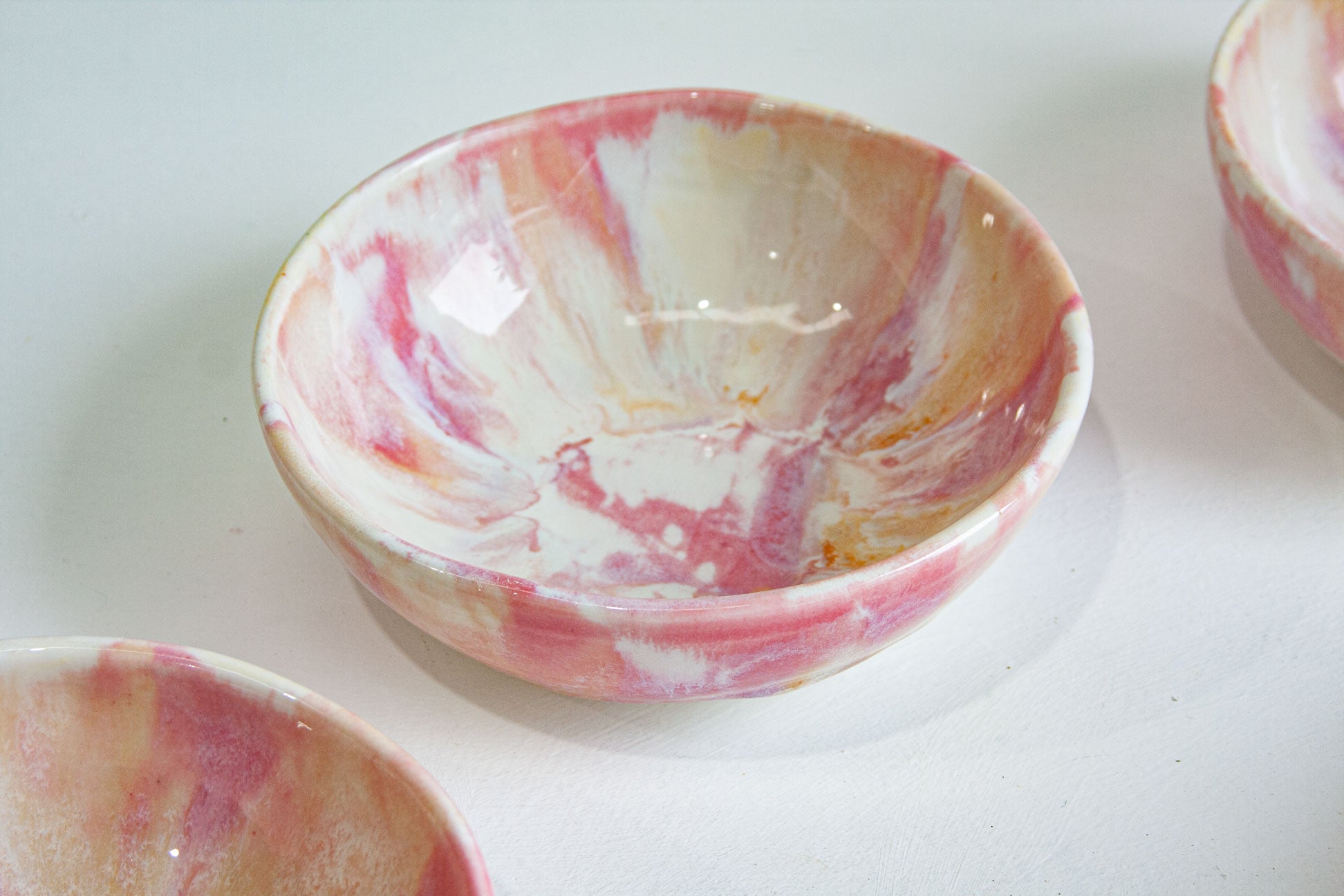 A handmade ceramic bowl with a pink, white, and beige marbled pattern, shown on a light gray surface with other partial bowls visible at the edges.