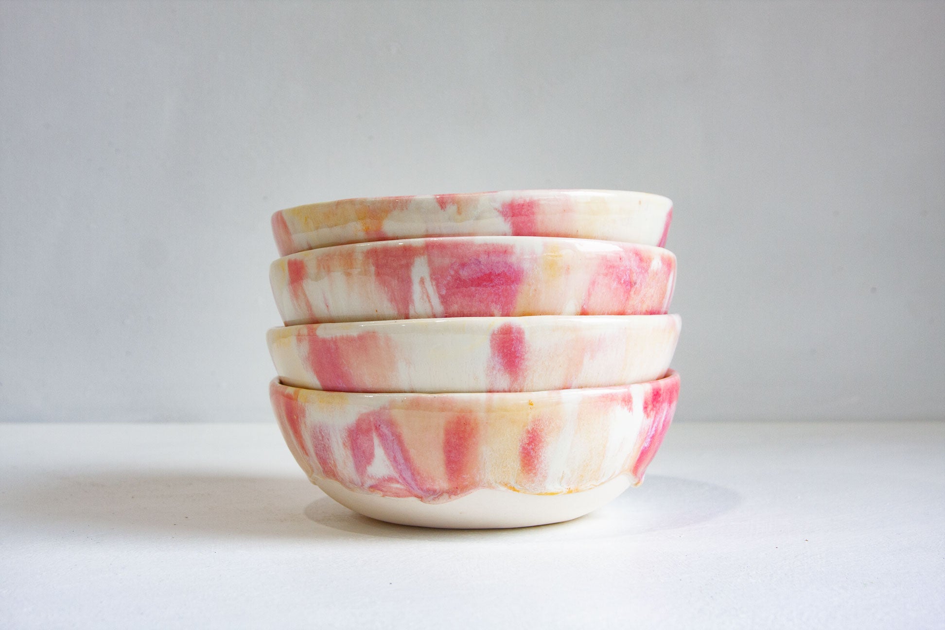 A stack of four small ceramic bowls with a marbled pattern of pink, peach, and cream on an off-white base, set against a neutral light gray background.