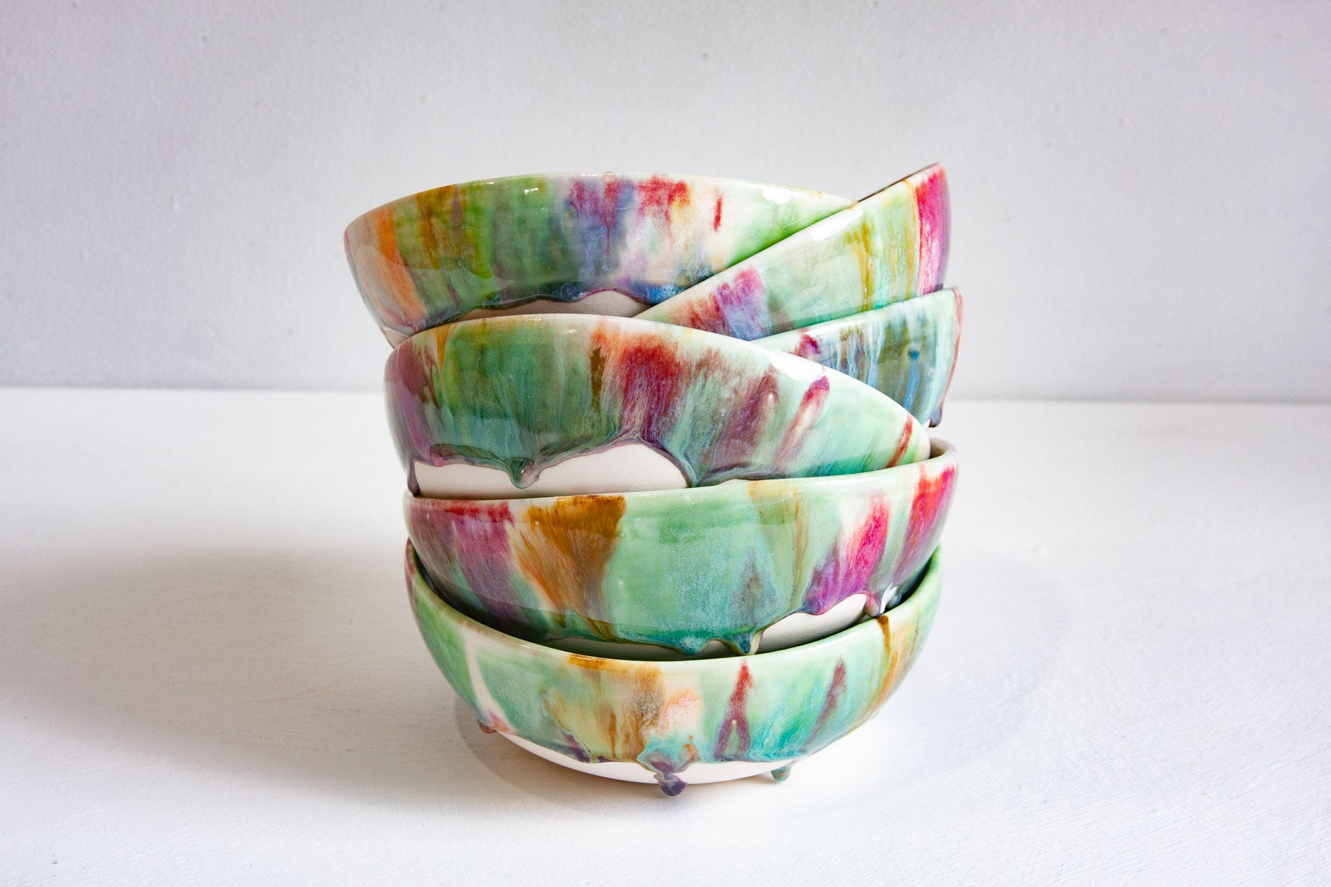A stack of four handmade ceramic bowls with a vibrant, multicolored marbled glaze featuring shades of green, pink, orange, and blue on a white base, set against a plain light gray background.