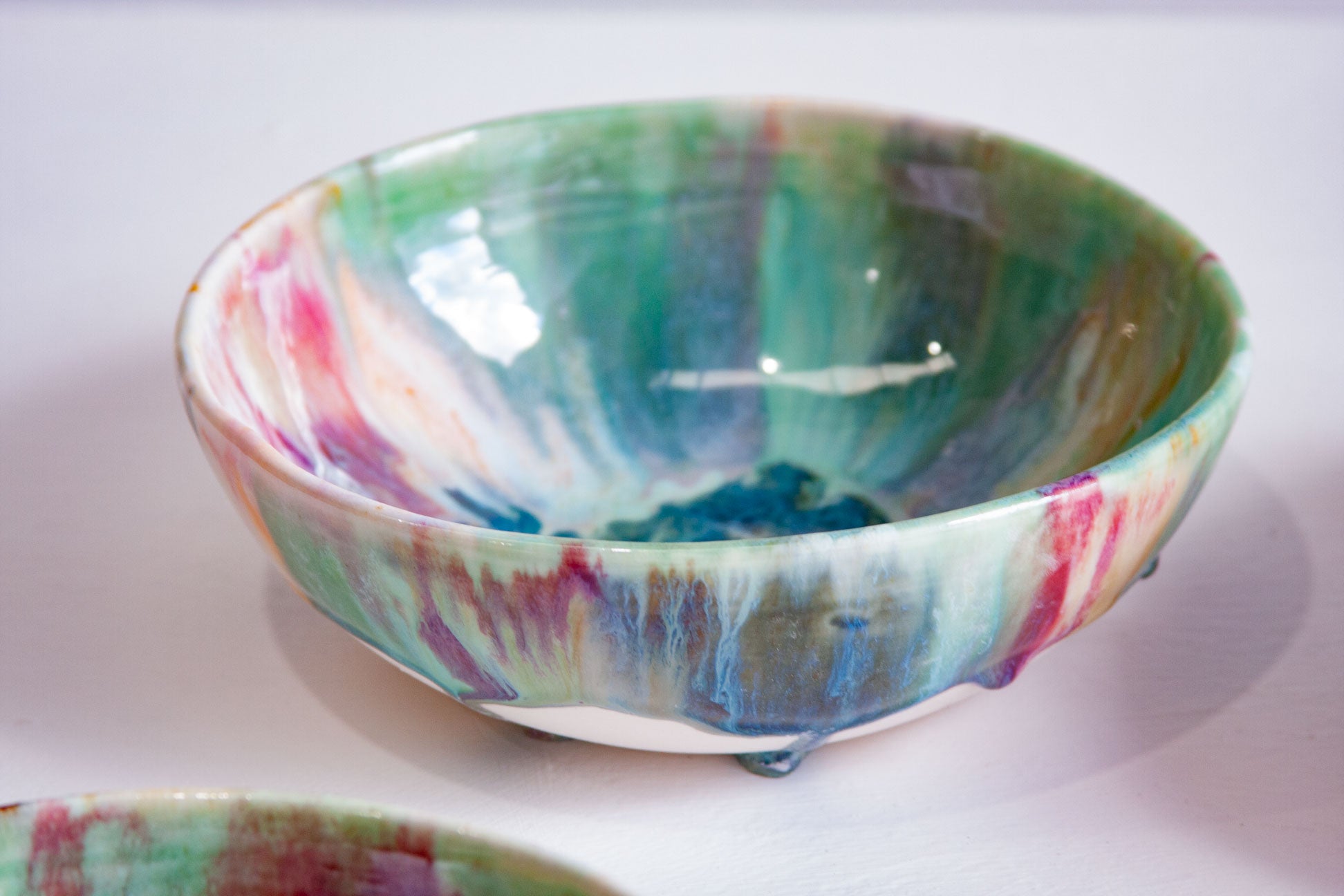 A handmade ceramic bowl with a marbled, drip-glazed finish in shades of green, blue, pink, and purple, shown on a white surface.