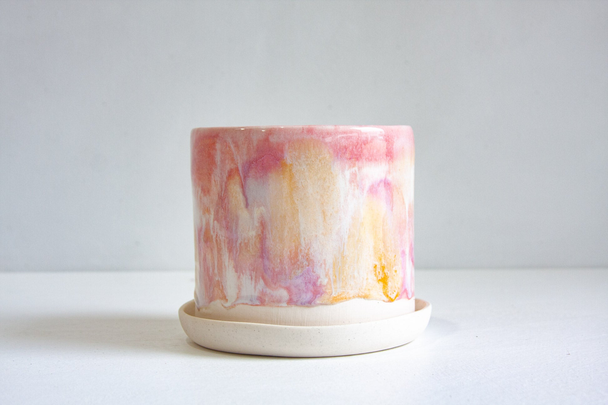 A handmade ceramic planter with a marbled pink, orange, and white glaze, sitting on a matching shallow saucer against a neutral light gray background.