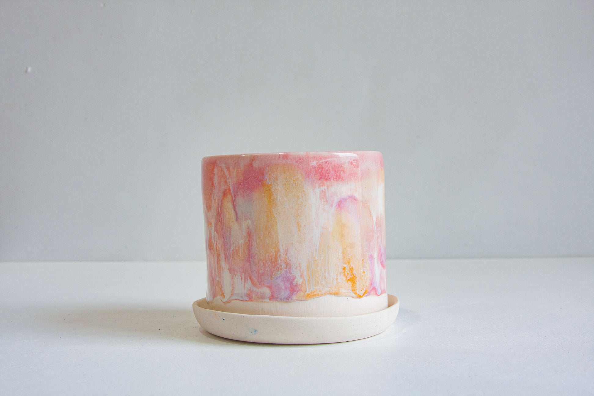 A handmade ceramic planter with a pink, orange, and white marbled pattern on the pot, resting on a matte off-white saucer against a neutral light gray background.