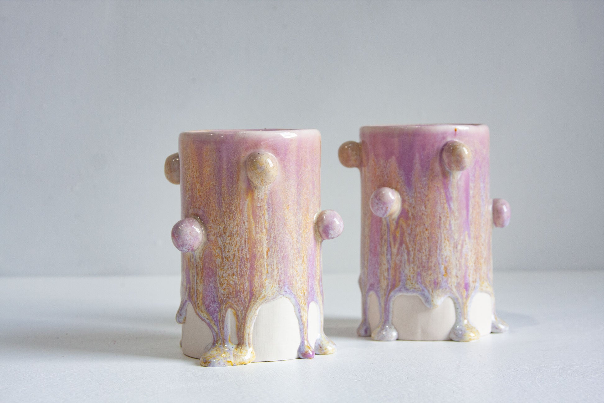Two small ceramic vases with a dripping pastel pink, purple, and gold glaze, standing on a white surface against a light grey background.
