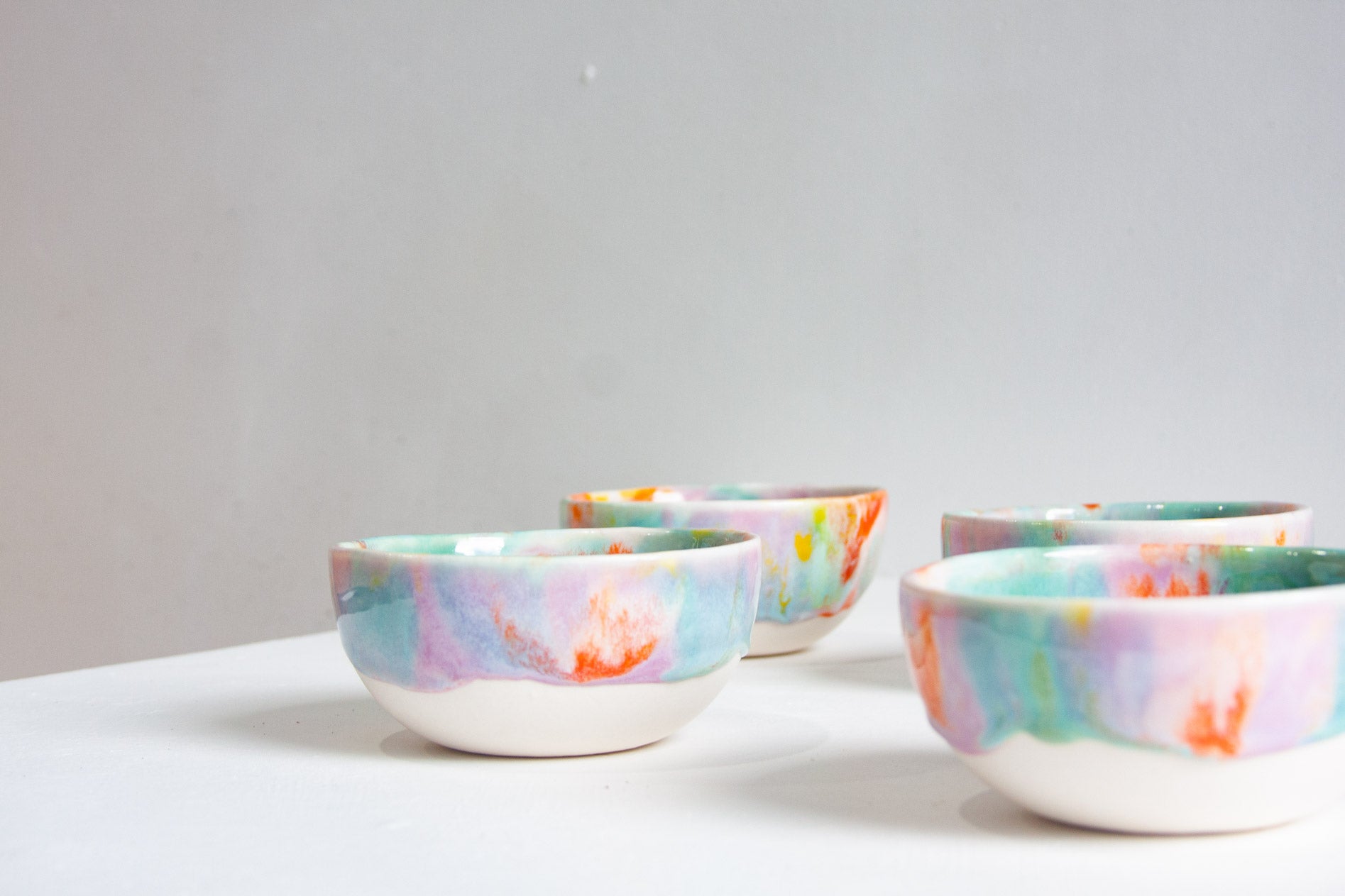 Four small ceramic bowls with a colorful pastel marbled pattern in pink, blue, green, and orange on a white base, arranged on a white surface against a light gray background.