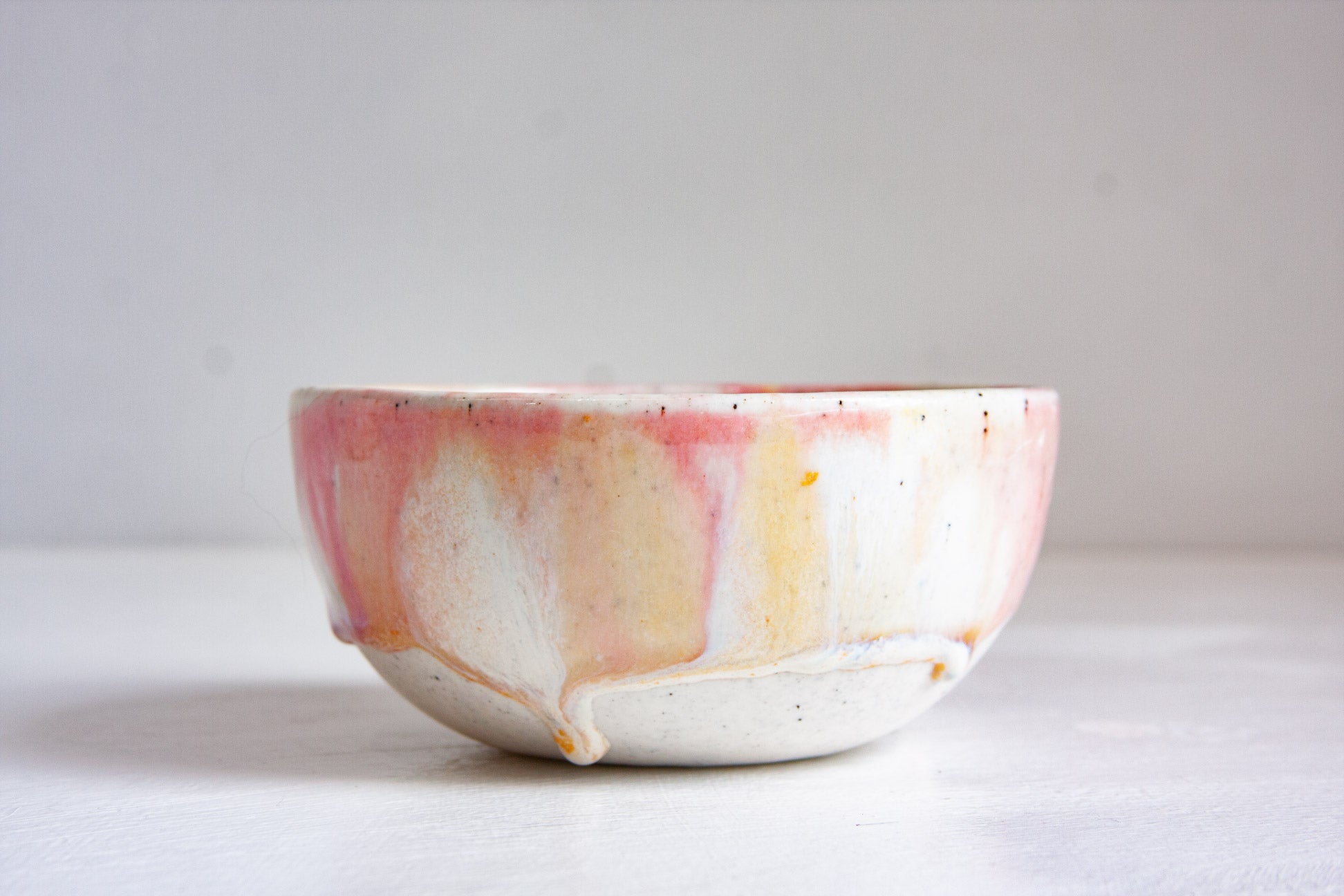A handmade ceramic bowl with a white base glaze and pink, yellow, and orange drip patterns on the exterior, set against a neutral light gray background.