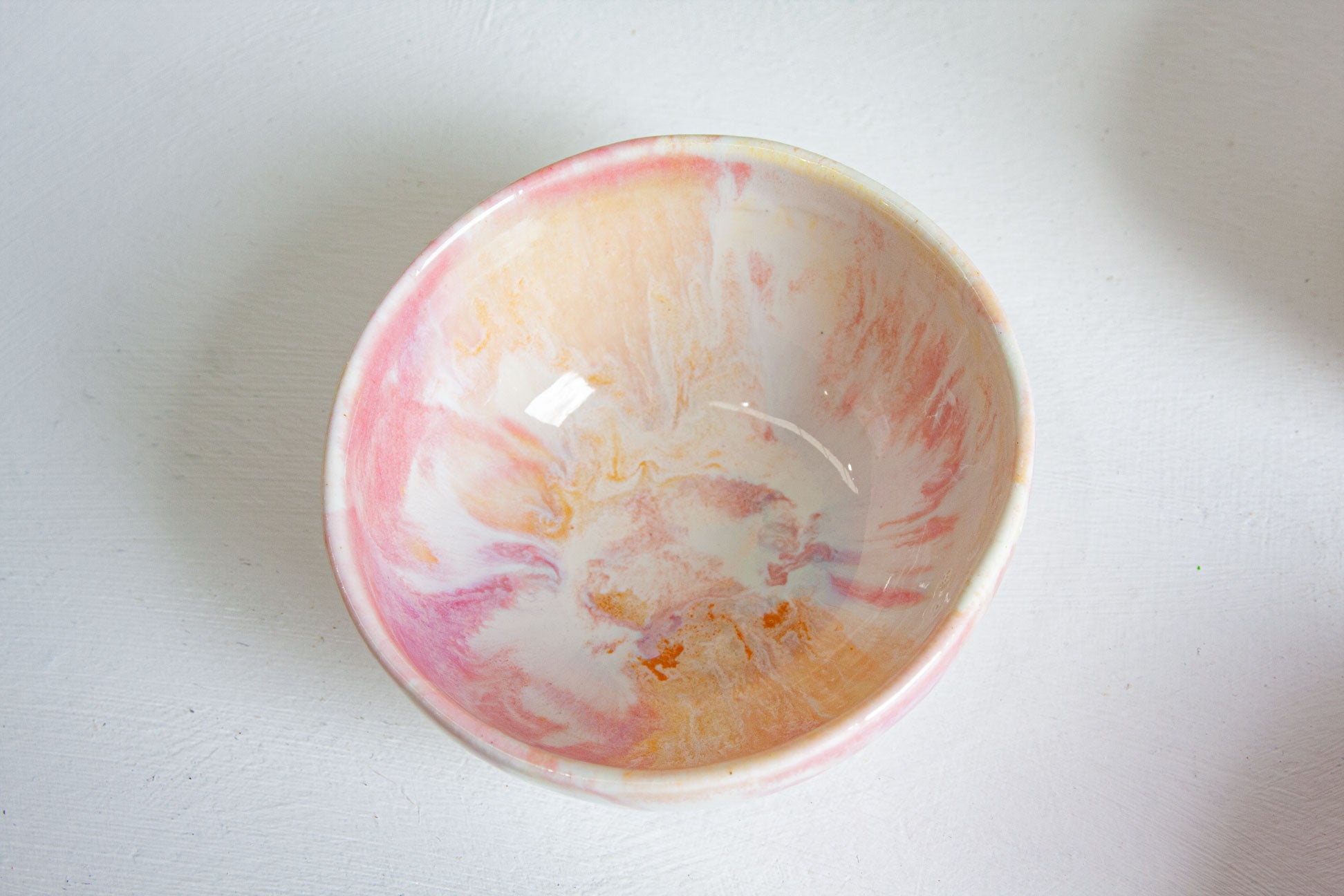 A handmade ceramic bowl with a marbled pattern of pink, orange, and white, photographed from a top-down angle on a white surface.