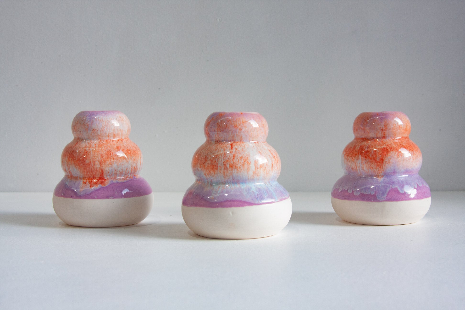Three small rounded ceramic vases with stacked organic shapes, featuring orange and purple marbled glazes on a white base, arranged on a white surface against a light grey background.