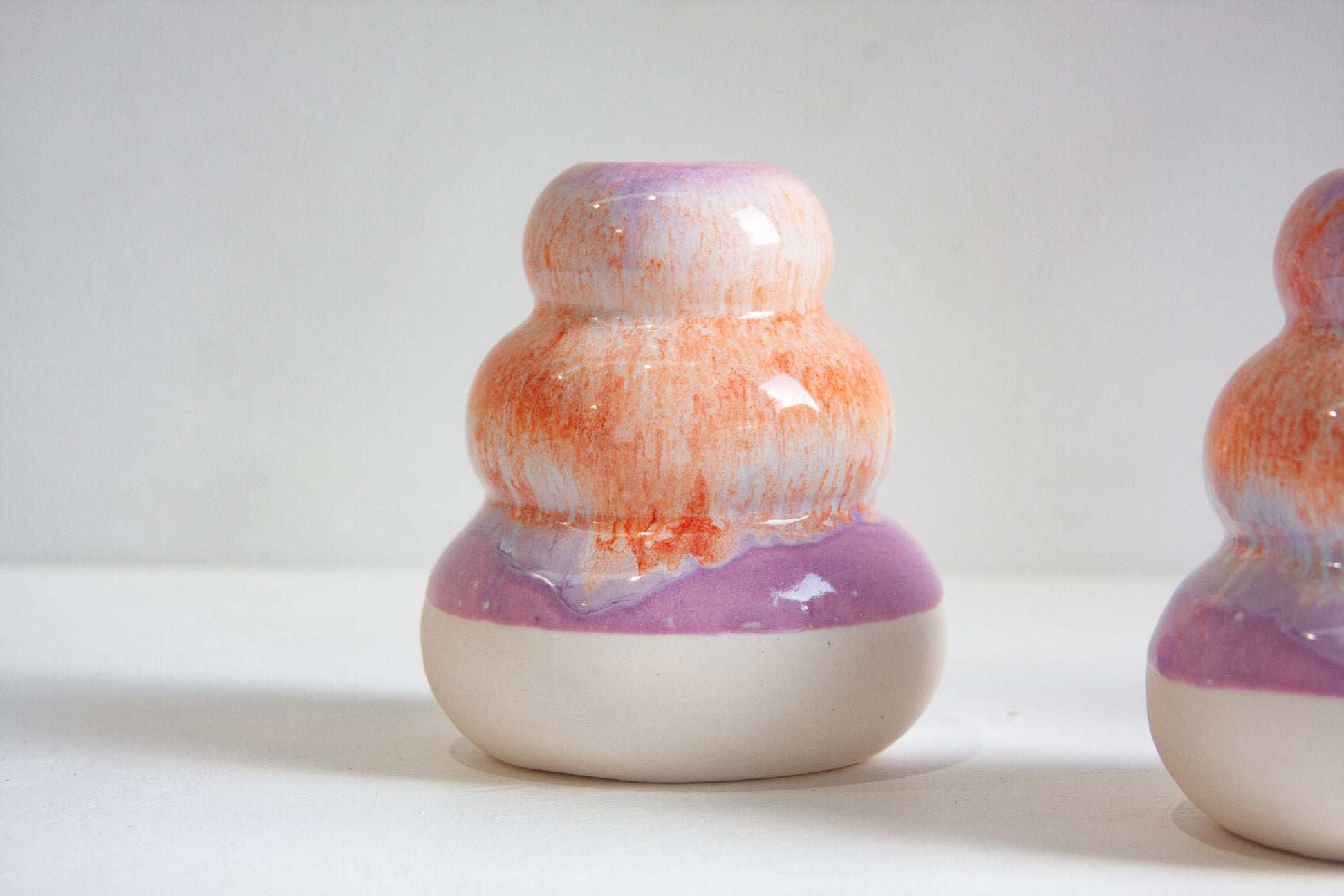 A small ceramic bud vase with a glossy glaze, featuring a white base, a purple band, and an orange-and-white marbled top section, shown against a plain light background.
