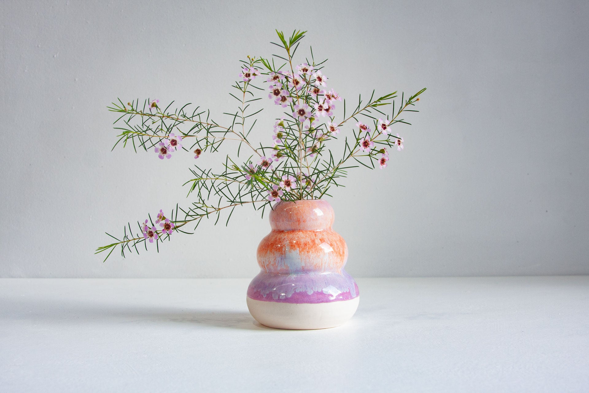 A small ceramic vase with a stacked, rounded shape in pastel orange, pink, purple, and white, holding delicate green sprigs with tiny pink flowers, set against a clean light grey background.