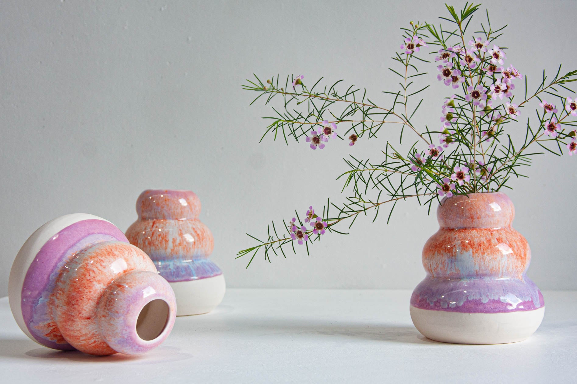 Three small ceramic bud vases with a marbled pastel orange, pink, purple, and white glaze are arranged on a white surface against a light gray wall. One vase is tipped on its side, while the other two stand upright, with delicate pink flowers placed in the rightmost vase.