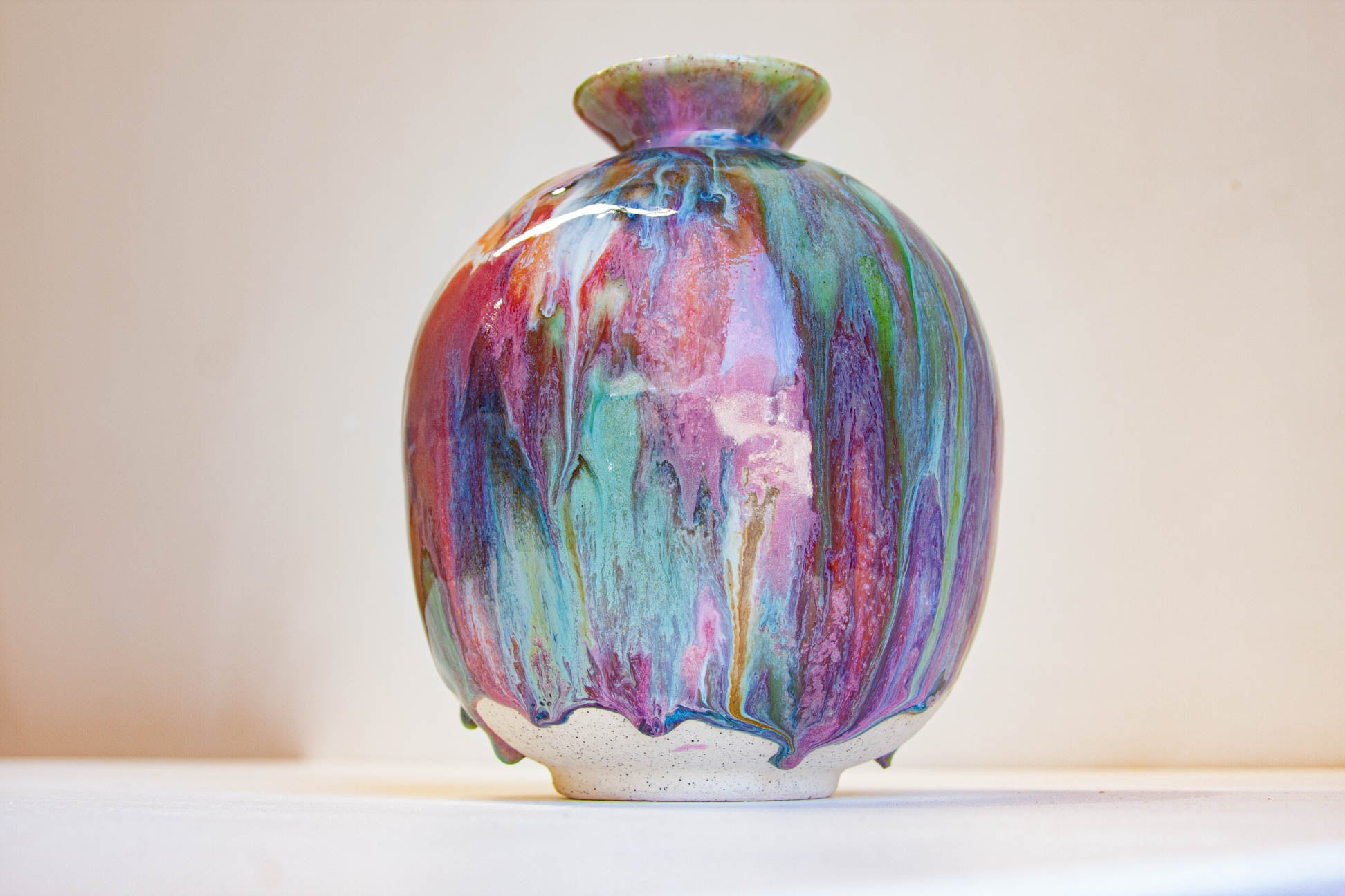 A small ceramic vase with a rounded body and narrow flared neck, decorated with a glossy multicolour drip glaze in pink, purple, blue, green, orange, and red tones, photographed against a plain light background.