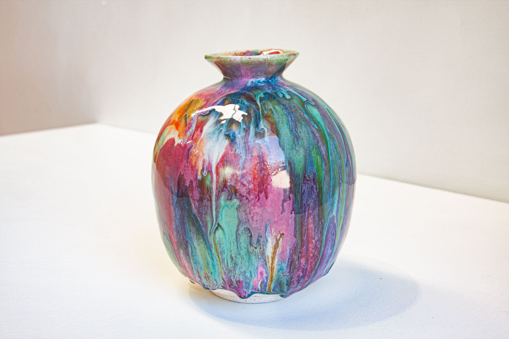 A small ceramic vase with a rounded body and short flared neck, covered in a glossy multicolor glaze with swirls of teal, purple, pink, orange, and green on a white surface against a light neutral background.